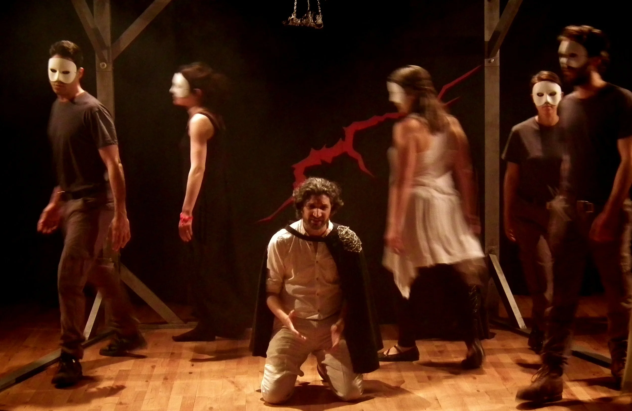 A theatrical stage scene with five performers wearing white masks. The central figure, kneeling and dressed in a white shirt and black cape, is gesturing with his hands. The background is black with a red lightning bolt symbol.
