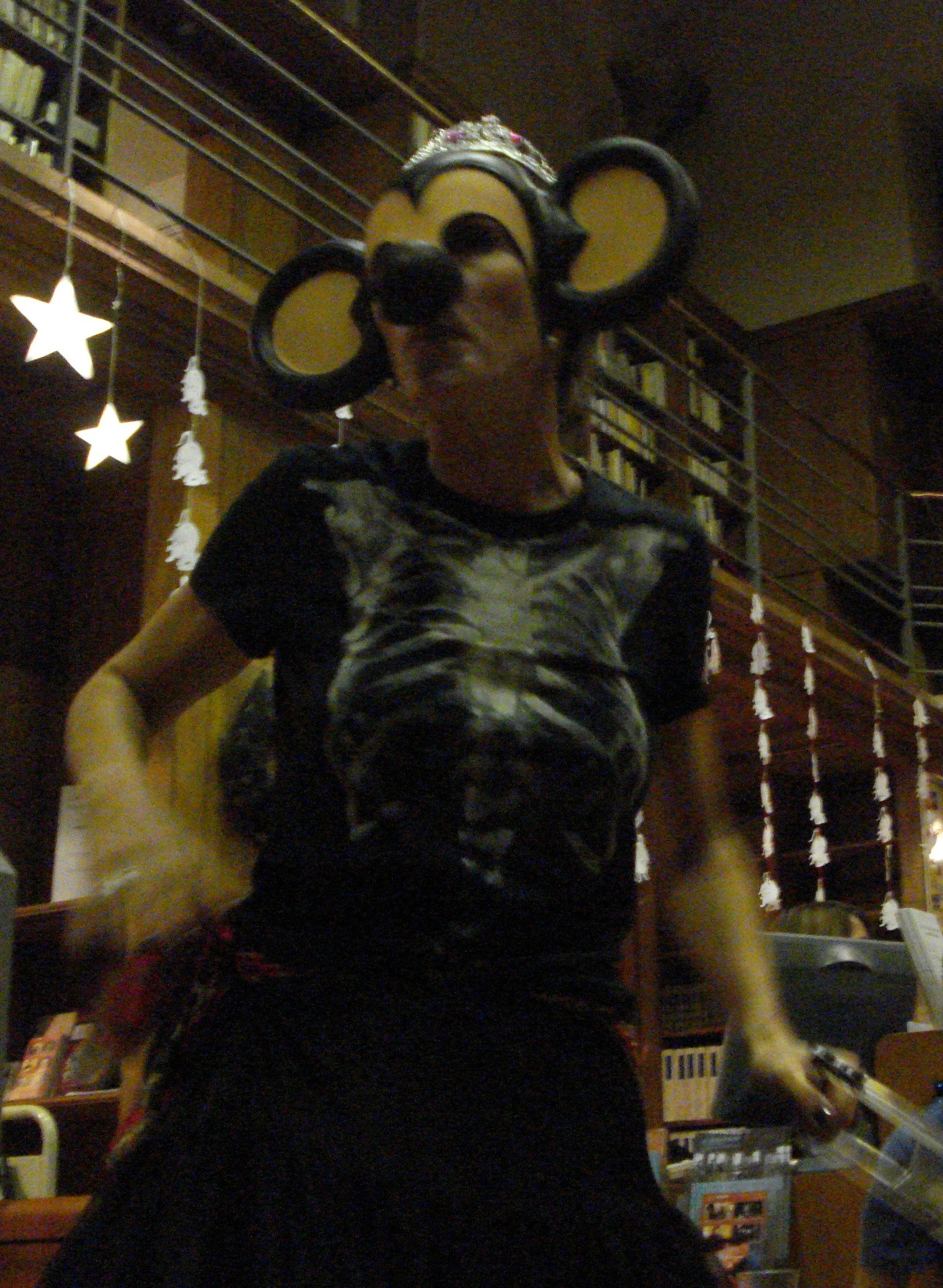 Une personne portant un masque de singe à grandes oreilles et une tiare, se tenant à l'intérieur d'une pièce faiblement éclairée, avec des décorations en forme d'étoiles et de Père Noël suspendues en arrière-plan.