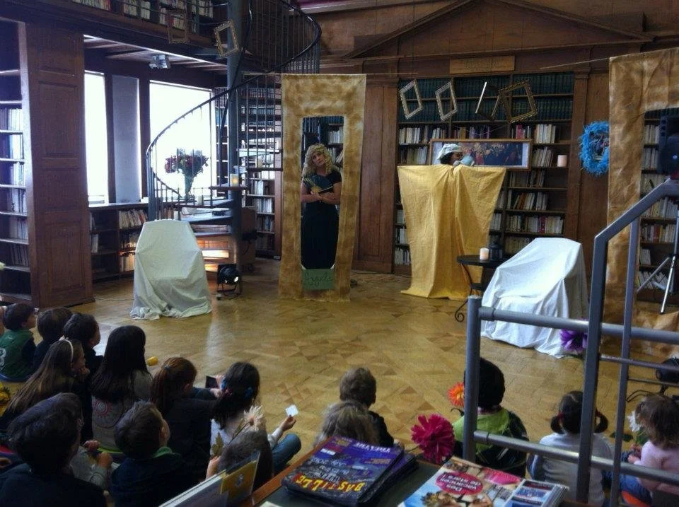 A show being performed in a library with children watching. There are stage props like chairs and mirrors, with bookshelves in the background.