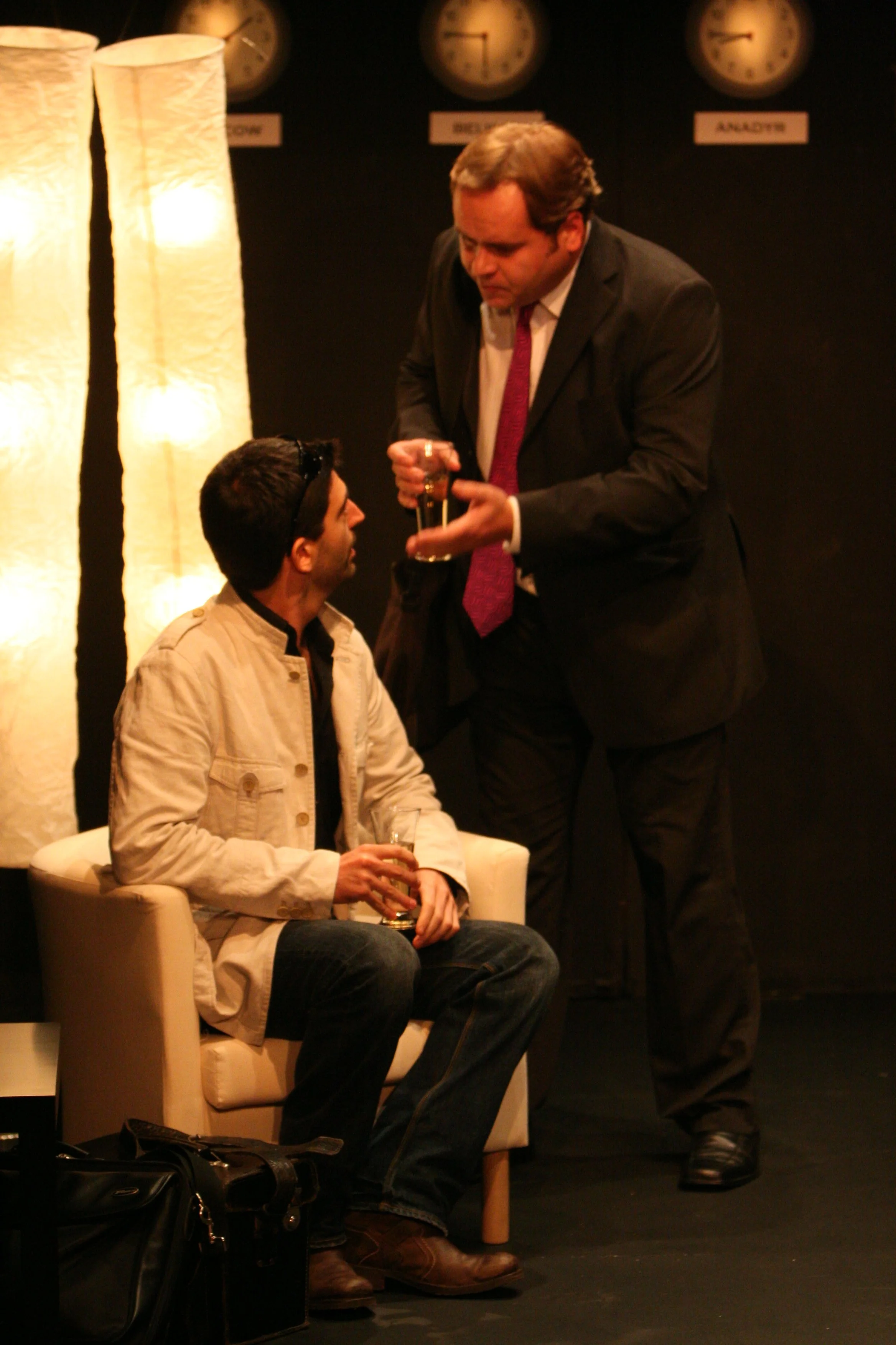 Two men onstage. One man wearing a suit is talking to another man sitting on a white chair. The man sitting is wearing a beige jacket and jeans, holding a glass. There are two tall lamps and three clocks on the wall behind them.