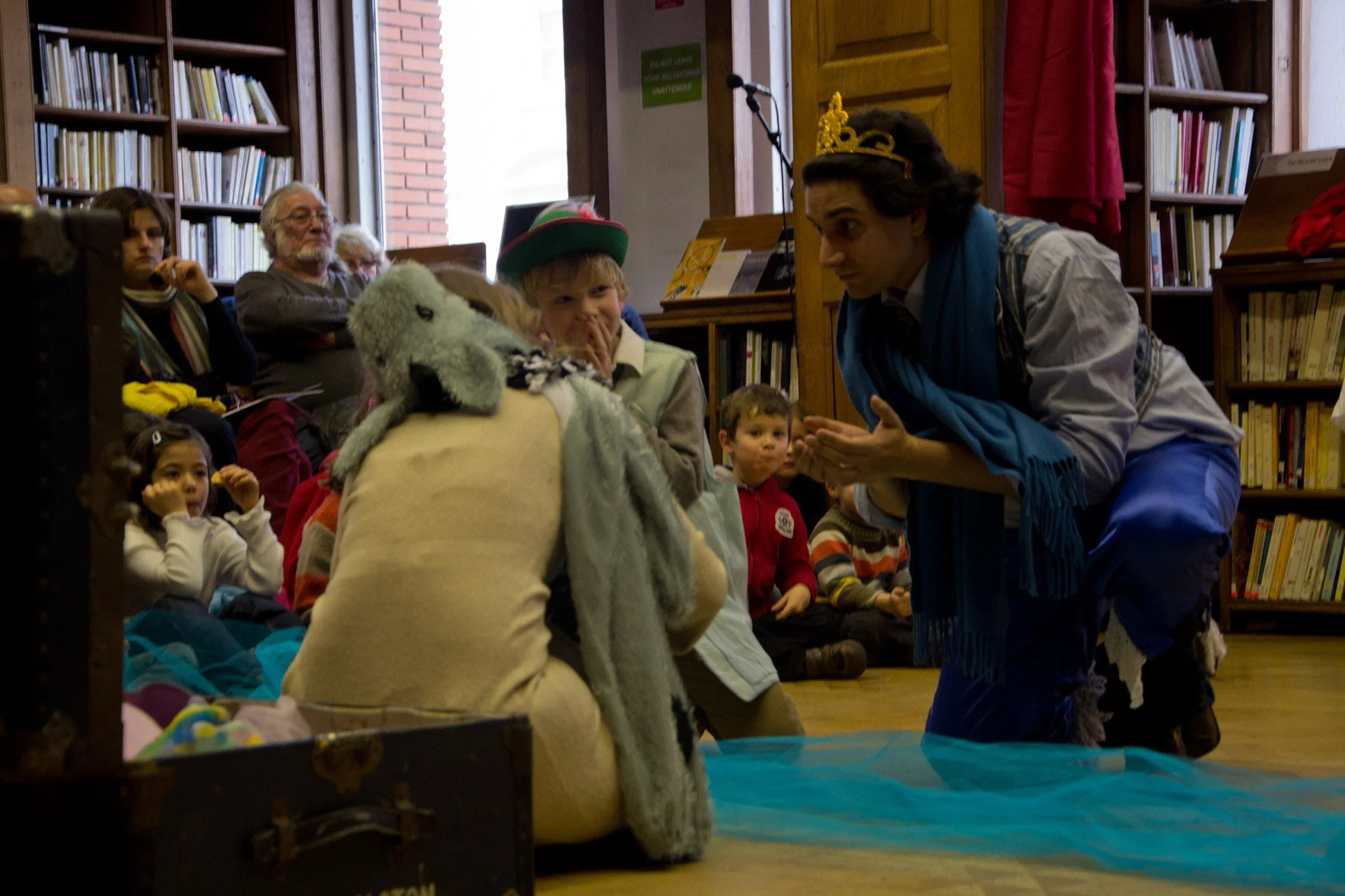 A man dressed as a king kneeling and talking to a child with a stuffed animal, surrounded by children and adults watching.
