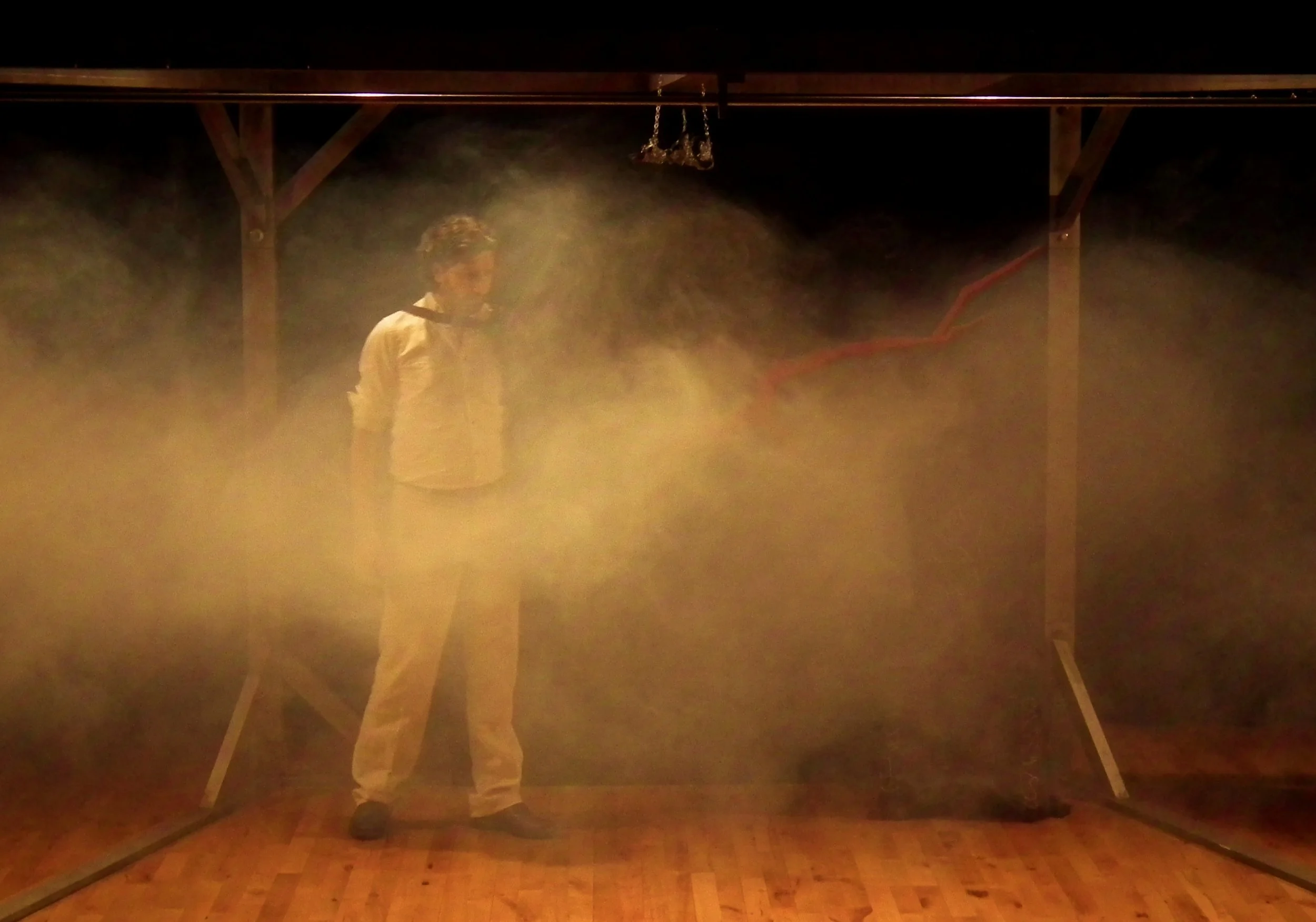 Person in white clothes standing inside a wooden frame, surrounded by thick yellowish fog, with a dark background.