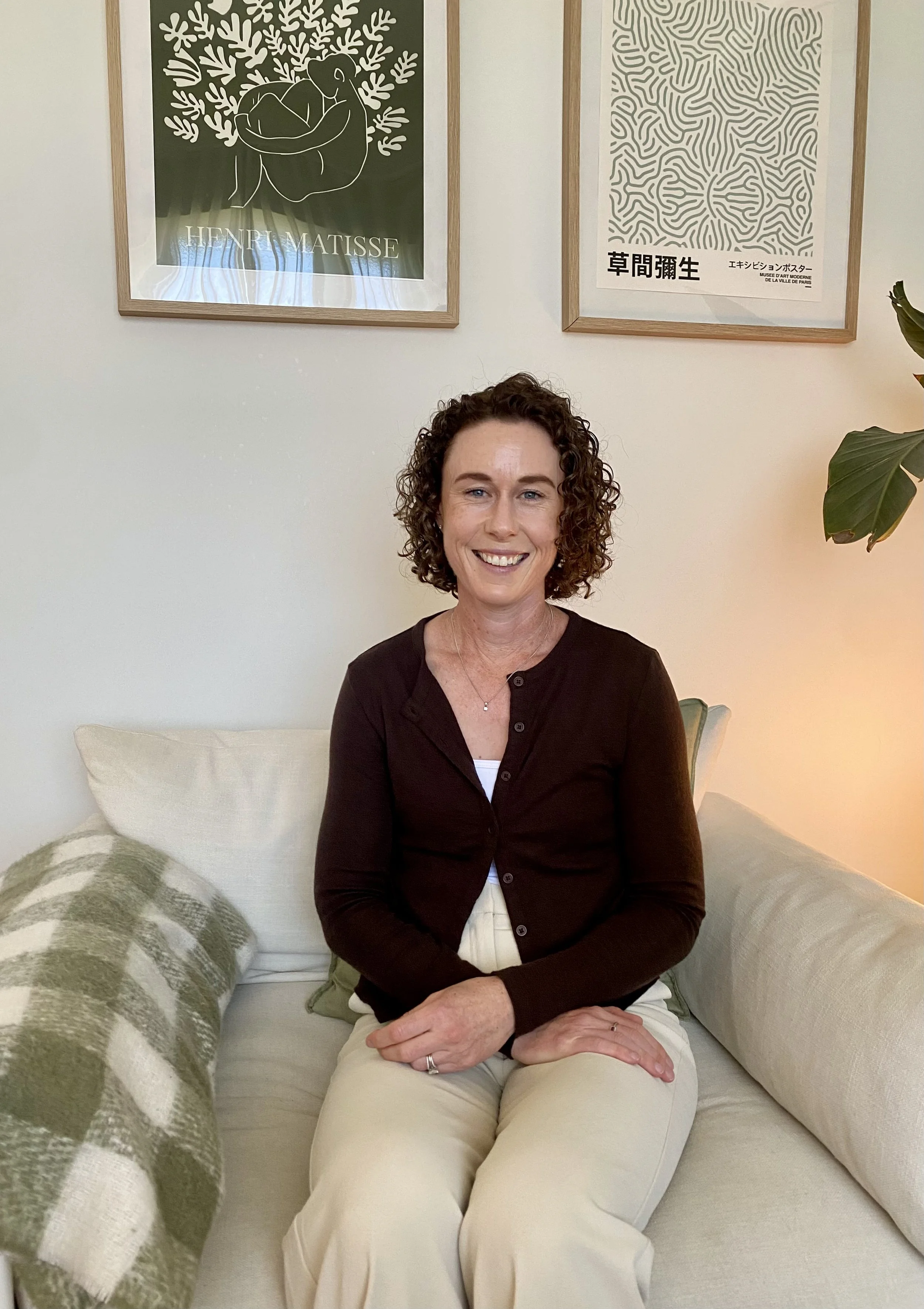 A woman with curly brown hair is sitting on a cream-colored sofa, smiling at the camera. She is wearing a black long-sleeve top and beige pants. Behind her on the wall are two framed art pieces, one featuring a minimalist image of a person sitting under a tree with the name 'Henri Matisse' at the bottom, and the other with abstract black and white patterns and Japanese text. A large green plant is partially visible on the right side.