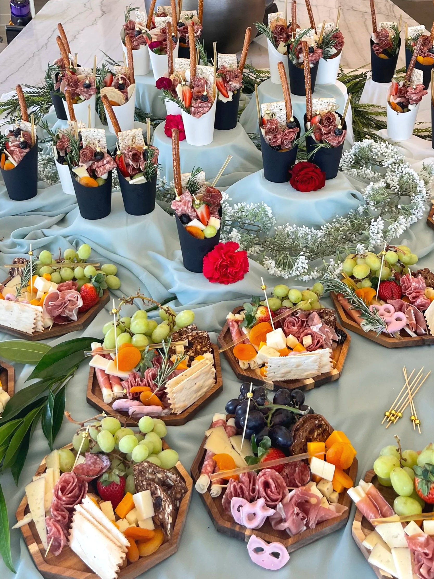 Mesa decorada con bandejas de queso, embutidos, uvas, fresas y frutas variadas, acompañadas de copas con aperitivos y decoraciones florales, ideal para una celebración.