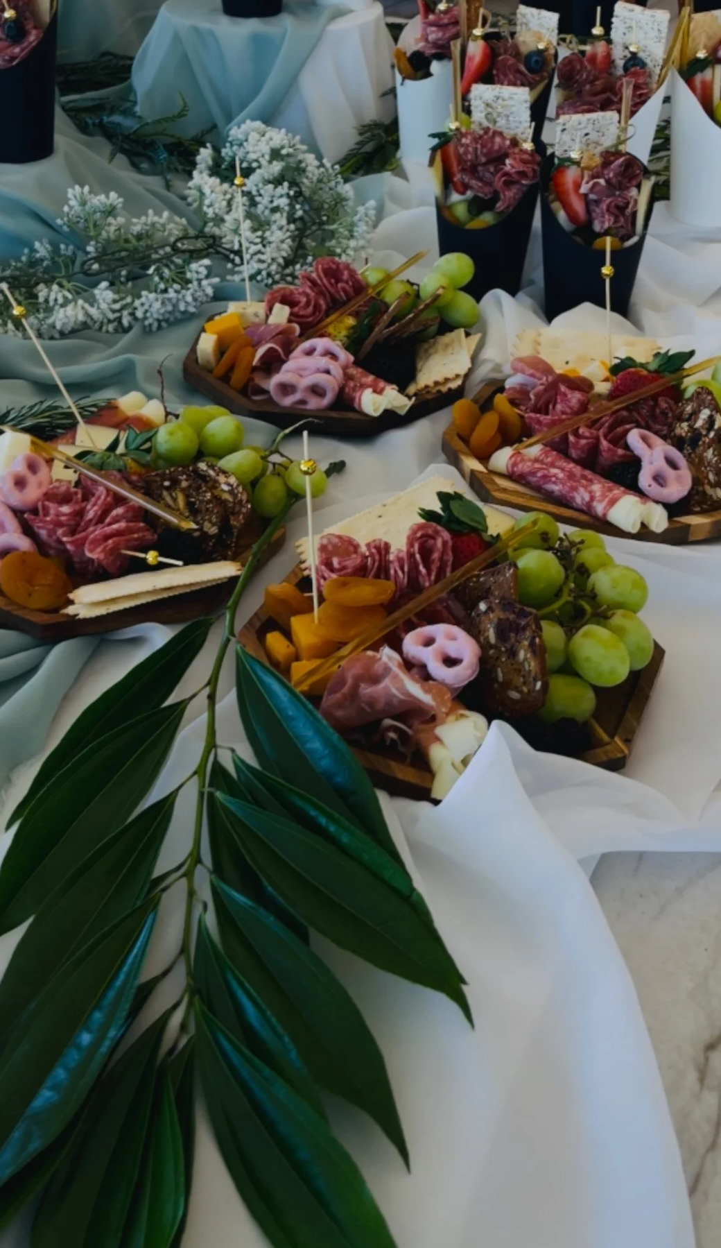 Tablas de charcutería con diferentes tipos de embutidos, quesos, uvas verdes, y frutas, decoradas con flores blancas y verdes, para una celebración o evento especial.