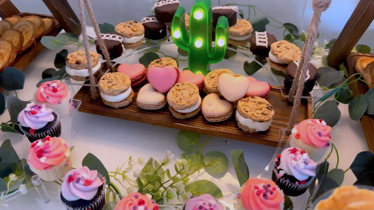 Mesa decorada con dulces, cupcakes con betún rosa y decoraciones en tonos pastel, galletas con Nutella, corazones de glaseado, y un cactus luminoso en medio, rodeada de hojas verdes y ramas.