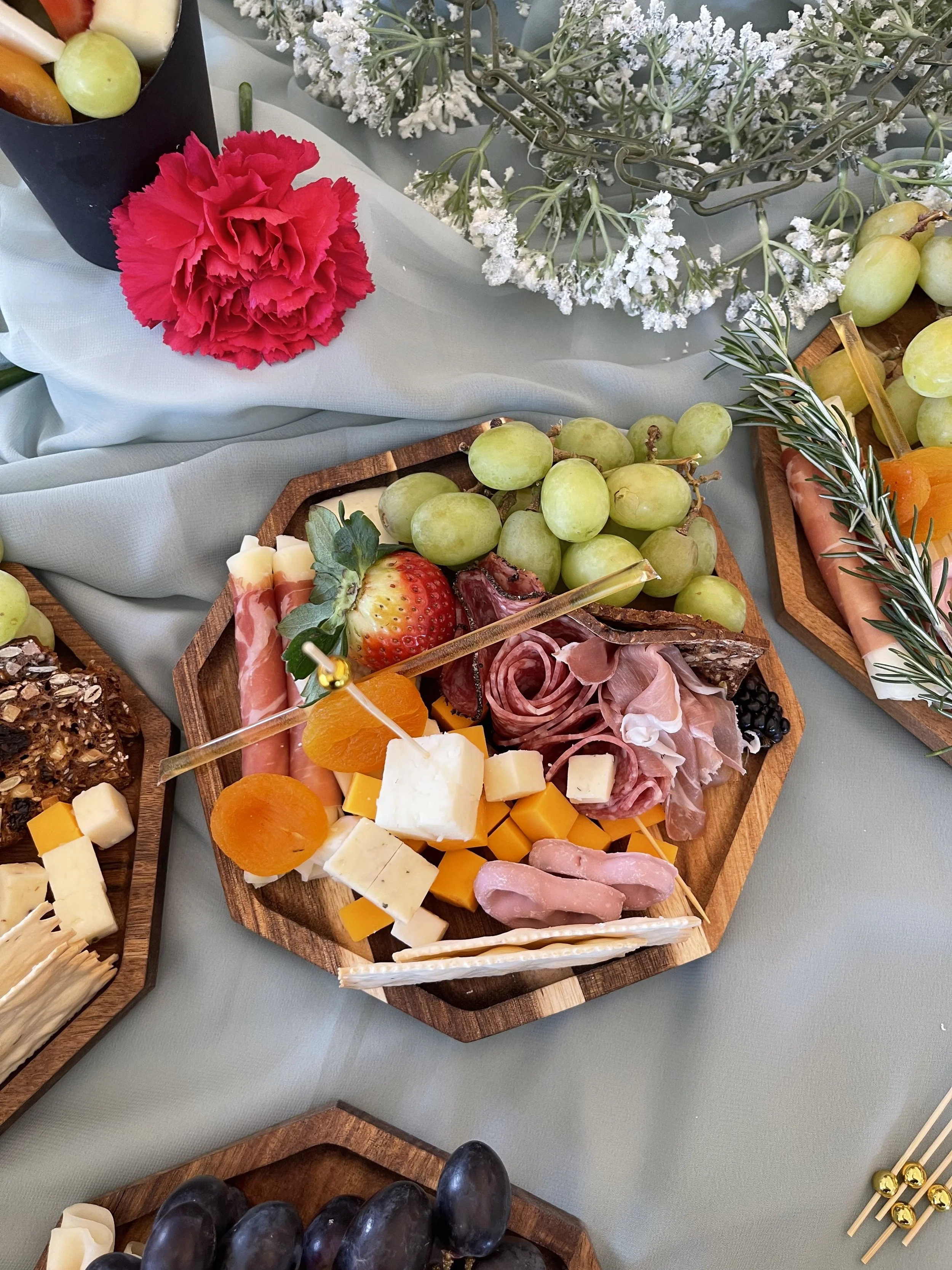 Tabla de aperitivos con uvas verdes, queso, jamón, fresa, mandarinas, y decoración de romero y flores blancas.