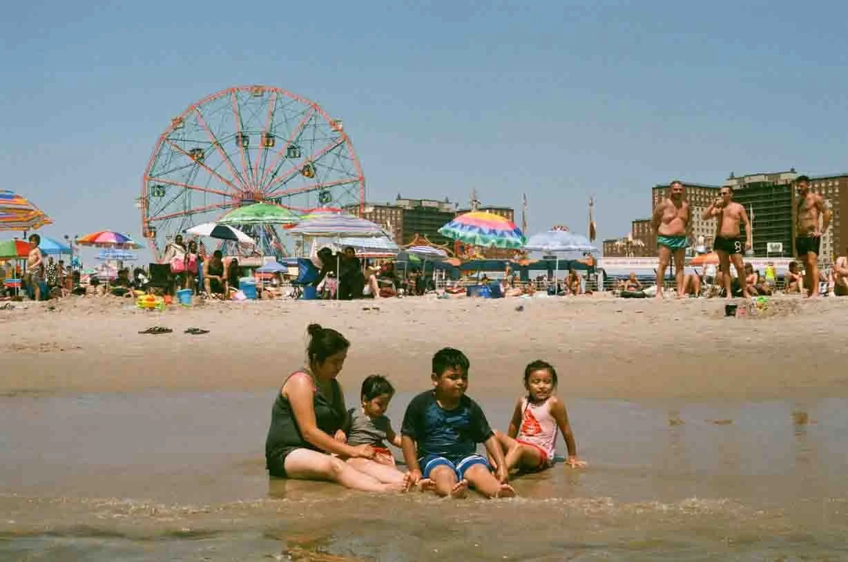 People enjoying a day at the beach with umbrellas, a Ferris wheel, and buildings in the background. In the foreground, a woman and three children sit in the water.