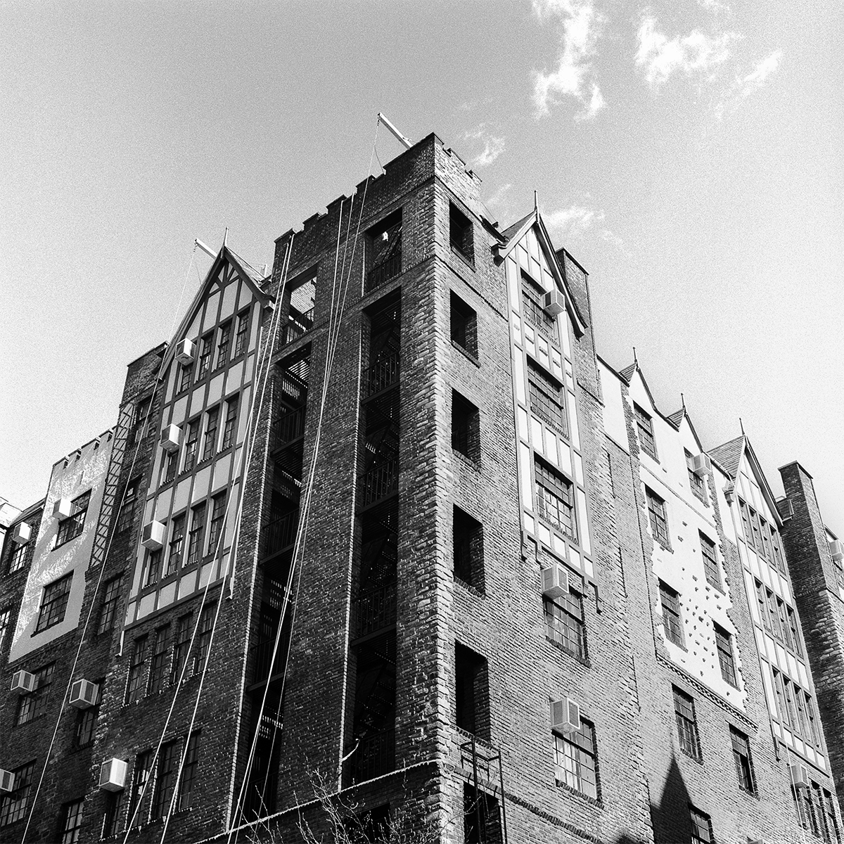 A black and white photo of a multi-story brick NYC building with pointed gable rooftops and several windows, some with air conditioning units, and fire escape stairs on the side.