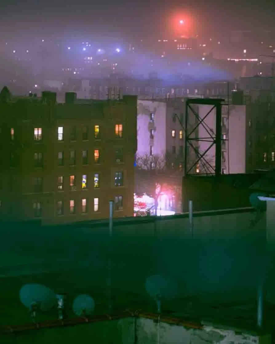 Nighttime cityscape with illuminated windows in buildings, a bright street light or firework in the sky, and city lights in the background, viewed from a rooftop.