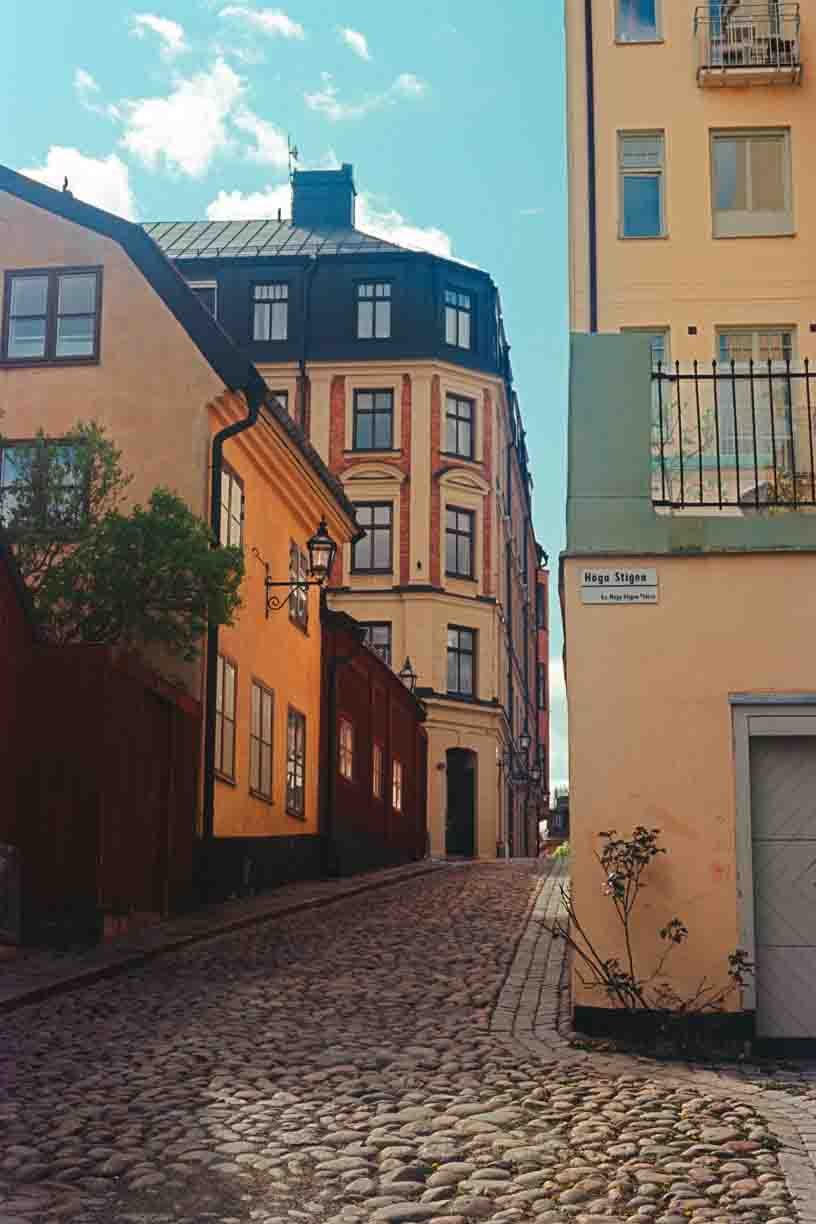 A narrow cobblestone street lined with colorful residential buildings, with blue sky and scattered clouds above.