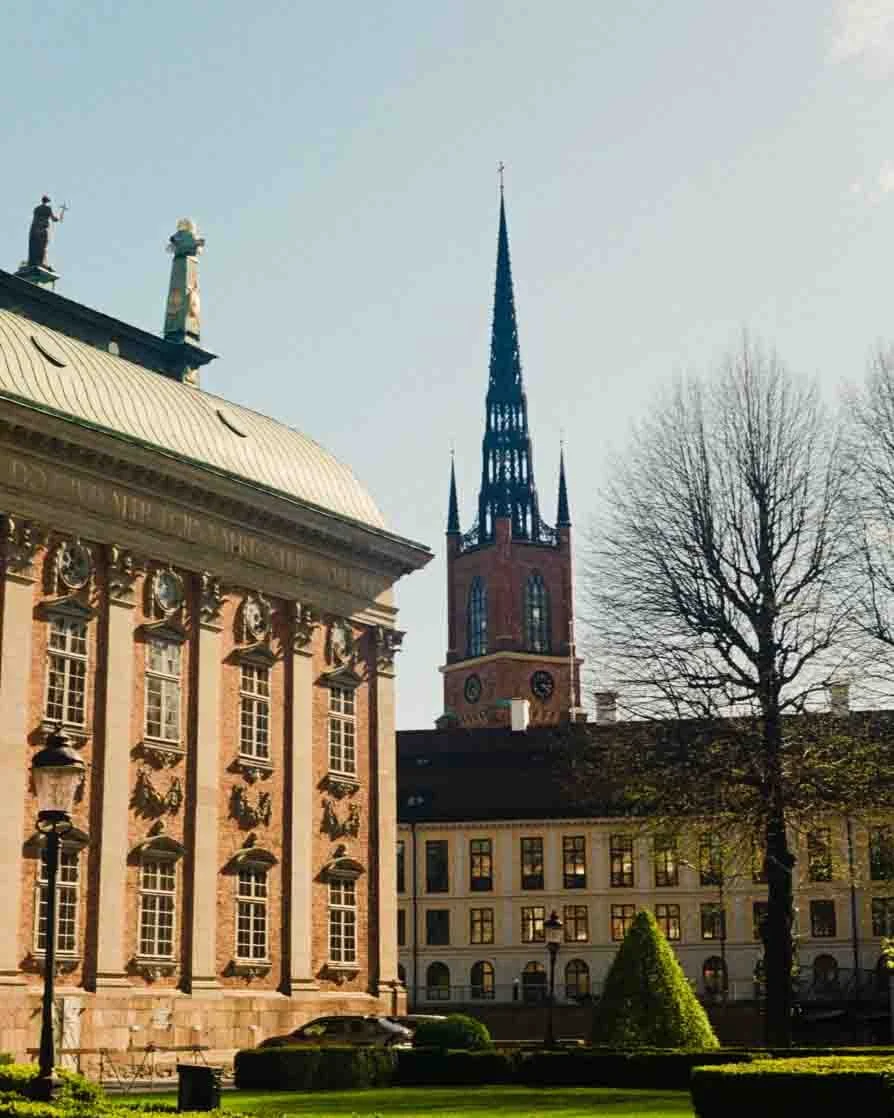 Historical European cityscape with a church steeple, classical building with ornate windows, leafless trees, and a well-kept lawn.