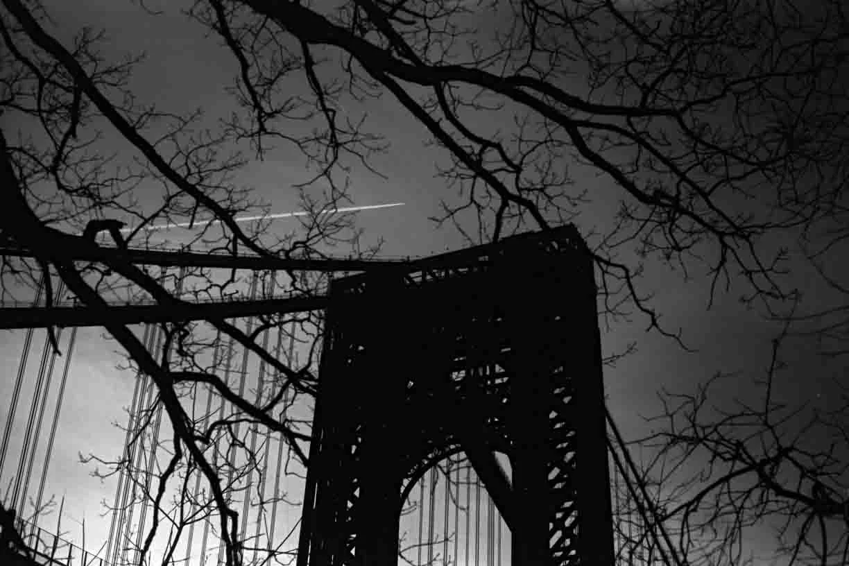 Silhouette of a bridge tower with bare tree branches against a cloudy night sky, with an airplane contrail visible.