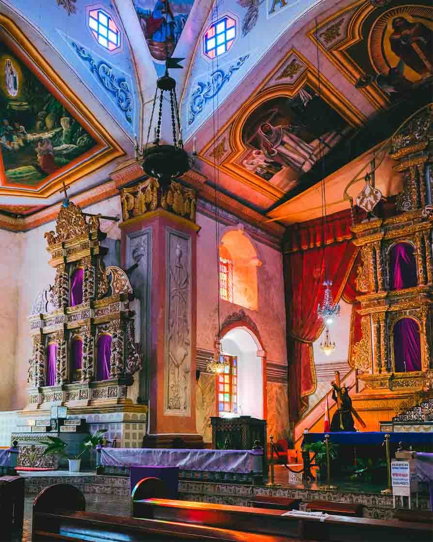 Interior of a church or cathedral with ornate gold decorations, purple curtains, stained glass windows, and a religious statue near the altar.