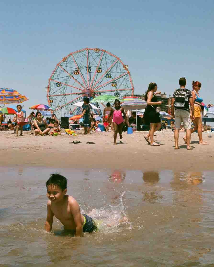 Children playing in the water on a busy beach with umbrellas, a Ferris wheel in the background, and people walking around and sitting on the sand.