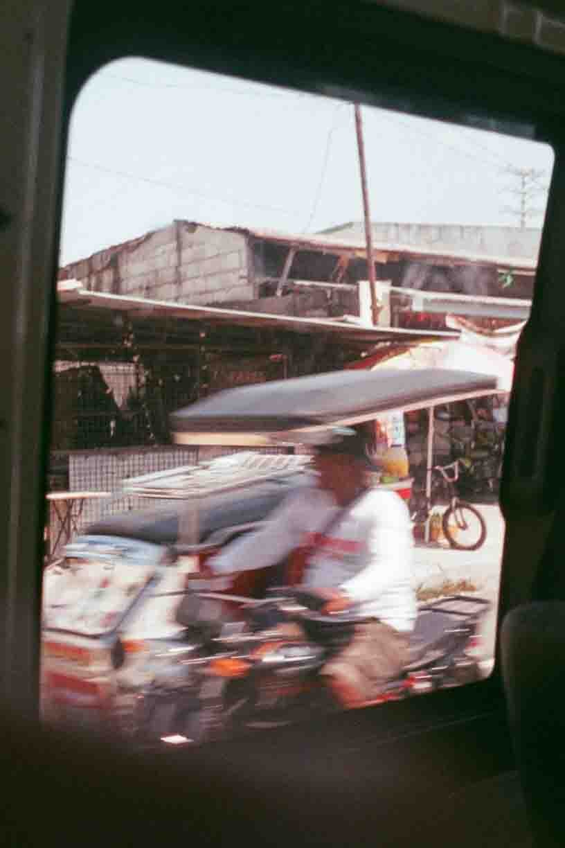 Photo taken through a vehicle window showing an urban street scene with a man in a white shirt and hat riding a motorcycle, other vehicles, and buildings in the background.