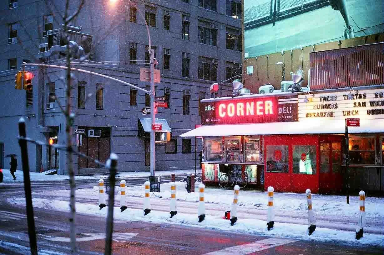 Nighttime scene of a snow-covered NYC street with a corner deli named 'The Corner.' The deli has a bright red neon sign, with a menu of food items displayed.  The street is lined with snow and safety barriers.