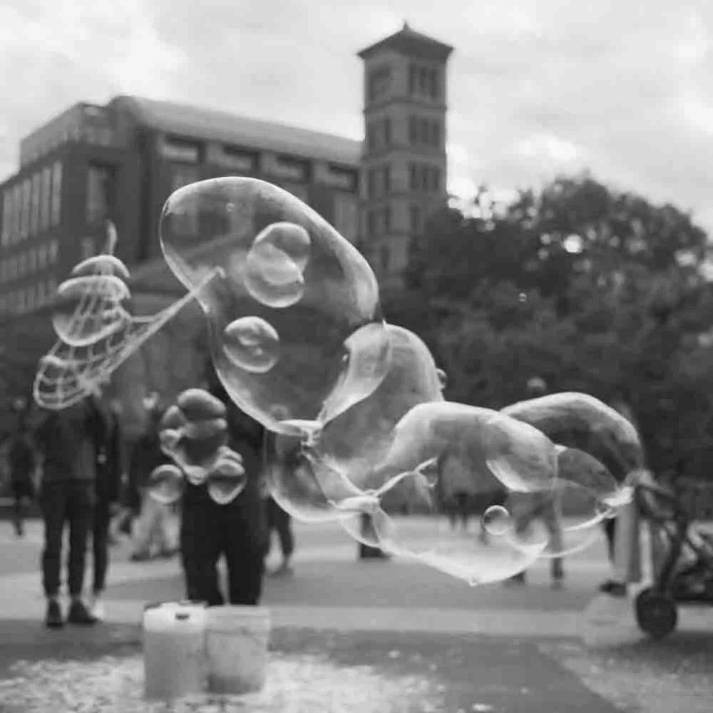 Bubbles floating in the air in front of a city square with buildings and people walking around.