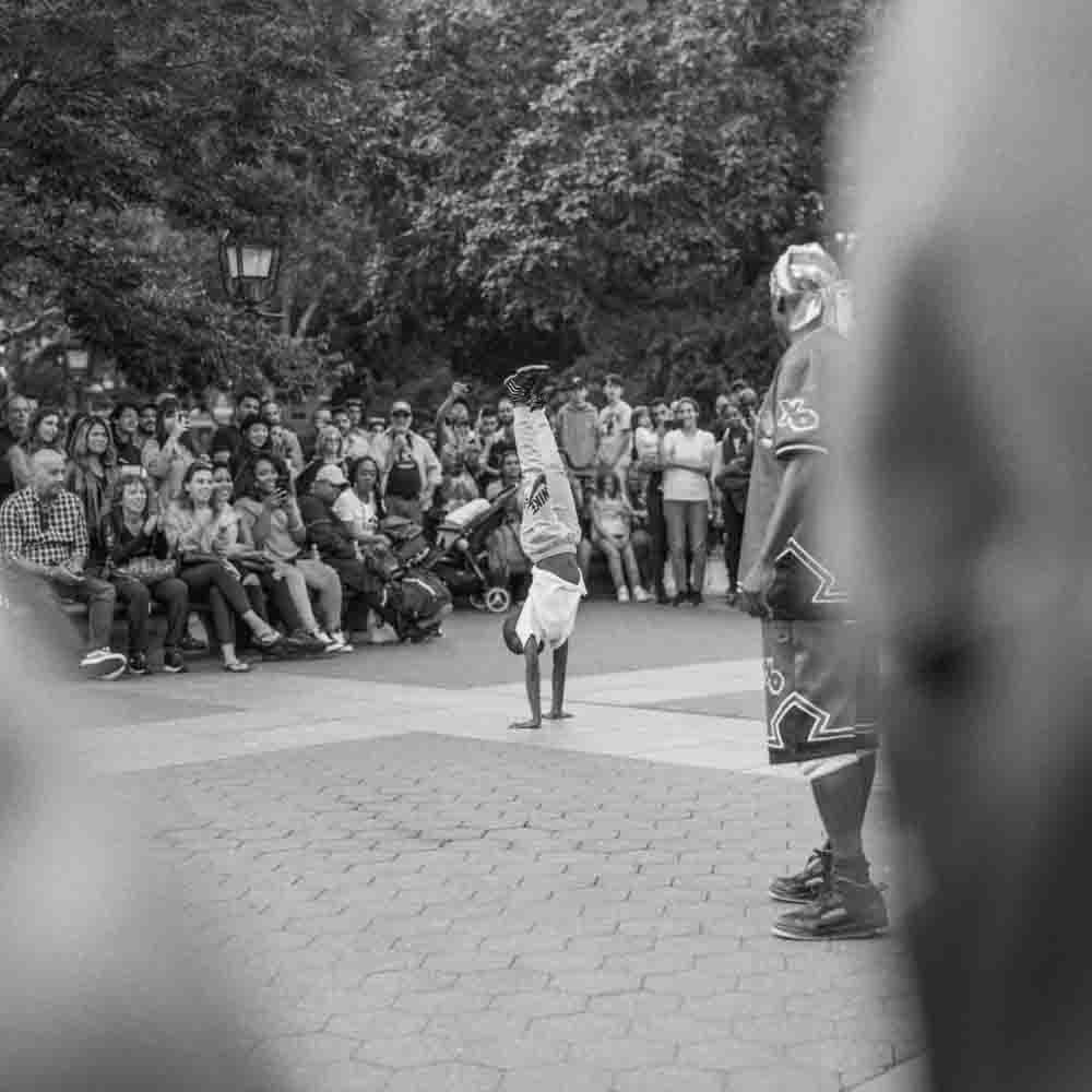 A street performer doing a handstand in front of a crowd at an outdoor park, with trees in the background.