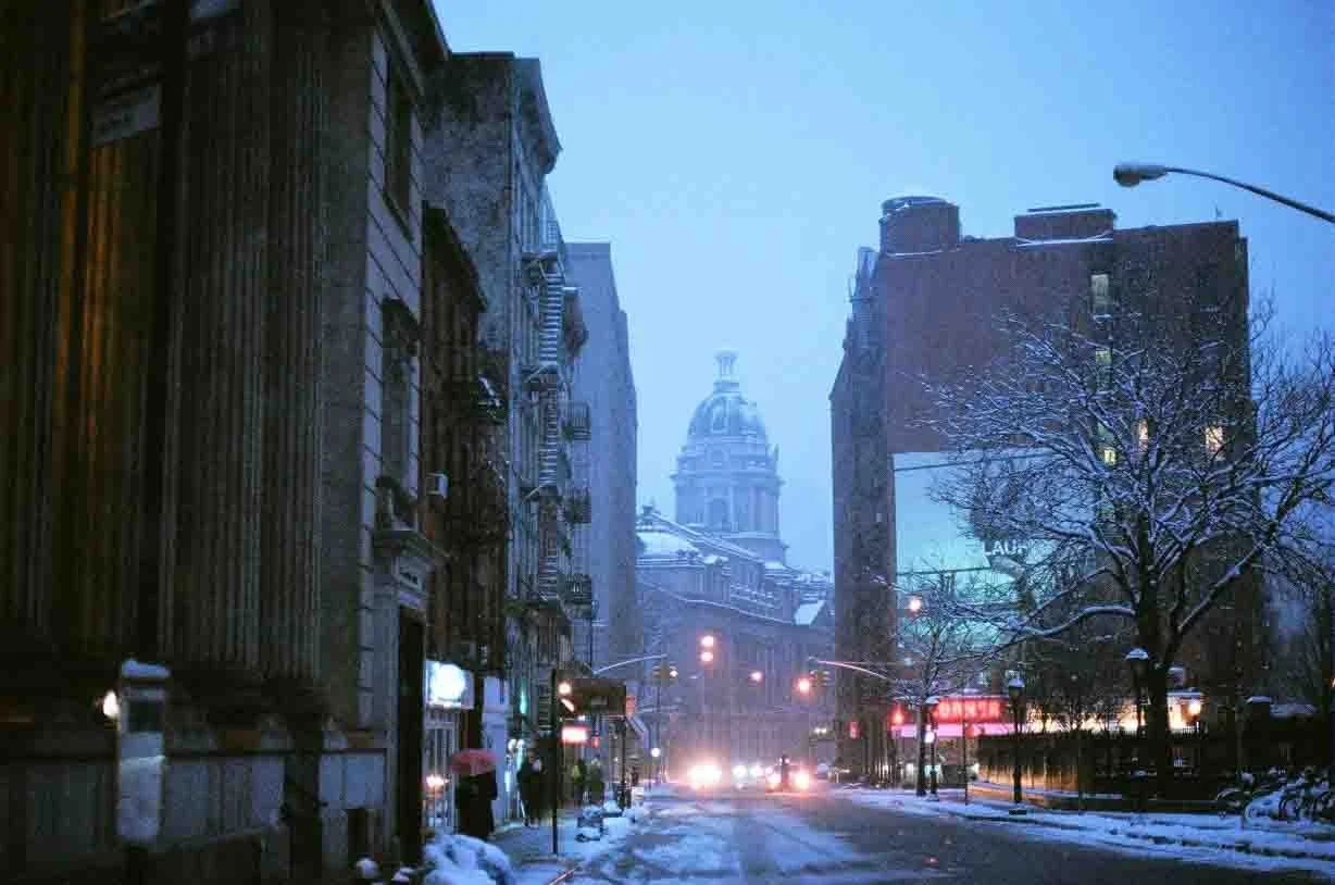 NYC street during winter snowfall with snow-covered sidewalks, leafless trees, and a historic domed building in the background under a blue evening sky.