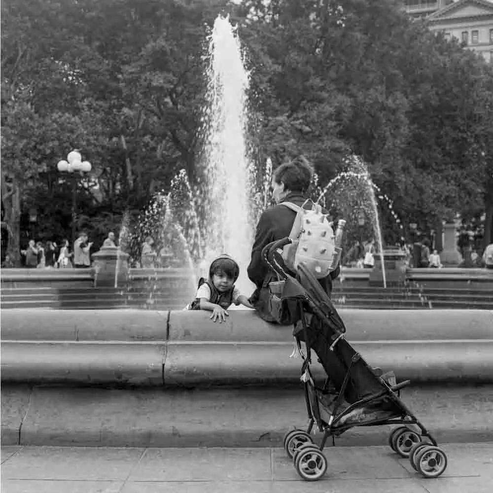 A person with a stroller and backpack sitting on the edge of a fountain, with a young child leaning over the fountain's ledge, in a park with trees and people in the background.
