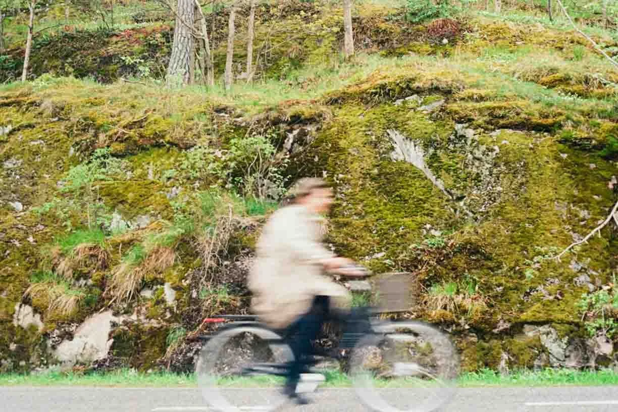 A person riding a bicycle on a road with a moss-covered hillside in the background.