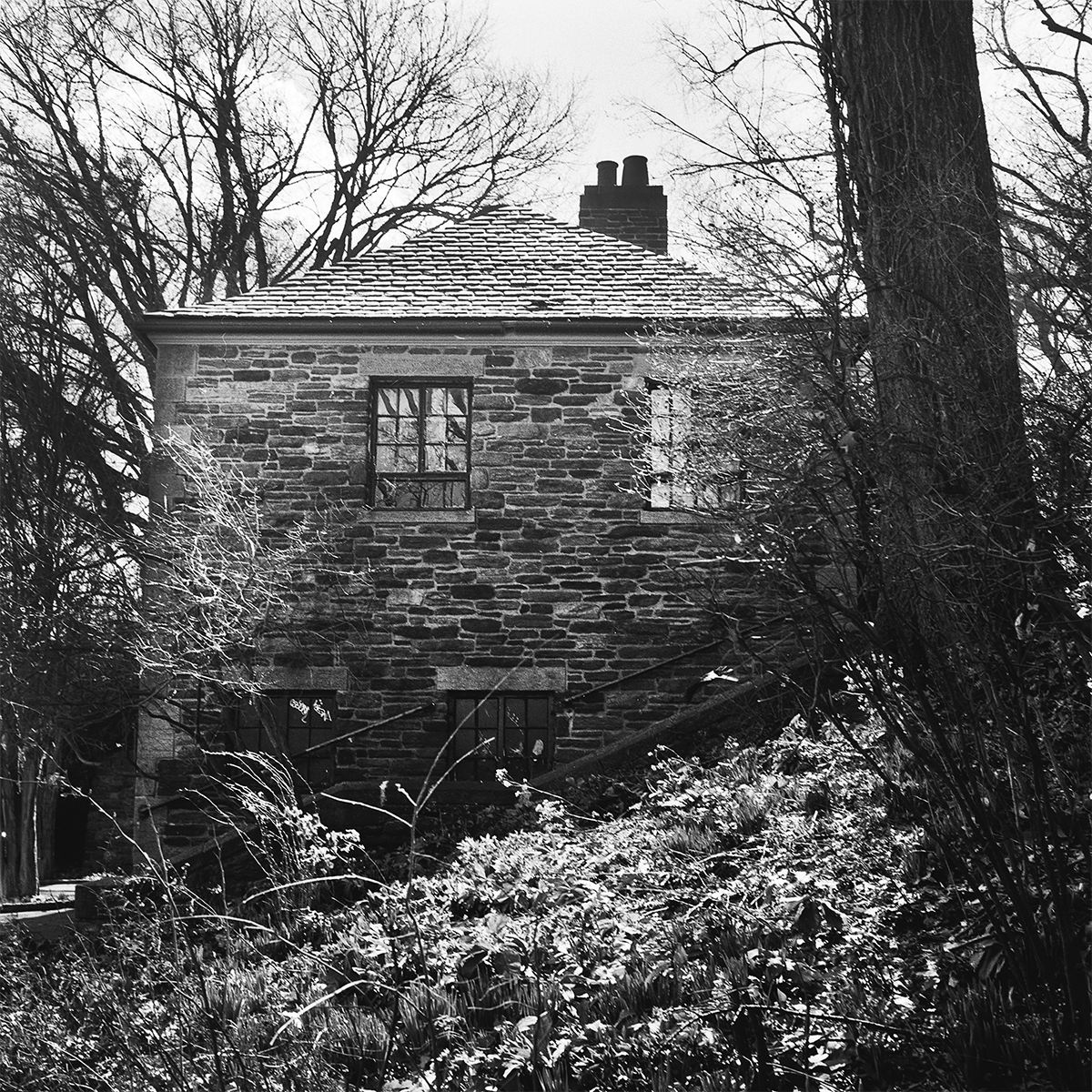 A black and white photo of an old stone house with a pitched roof, surrounded by leafless trees and overgrown shrubs.