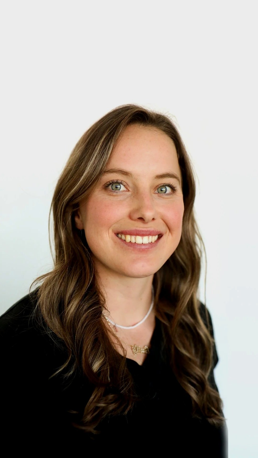 A smiling woman with long wavy brown hair, light blue eyes, and fair skin, wearing a black top and layered necklaces against a plain white background.
