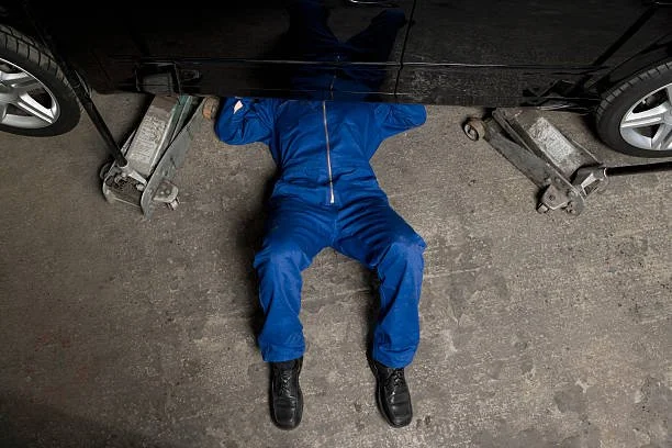 A person lying underneath a car, working on it from below, wearing blue coveralls and black shoes in an automotive garage.