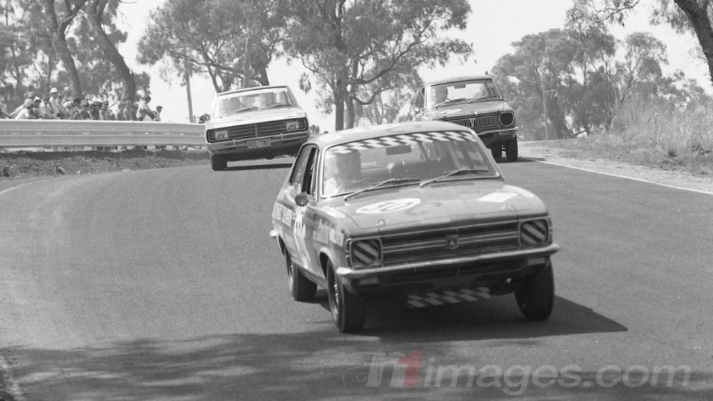 Vintage cars racing on a curving race track with spectators watching from the side, trees in the background.
