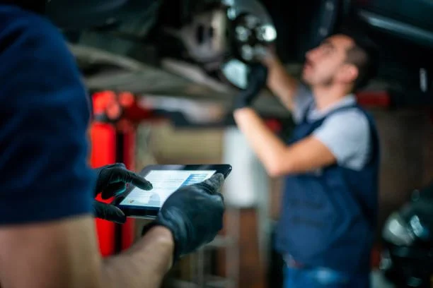 Mechanic working underneath a vehicle with another person using a tablet in a garage.