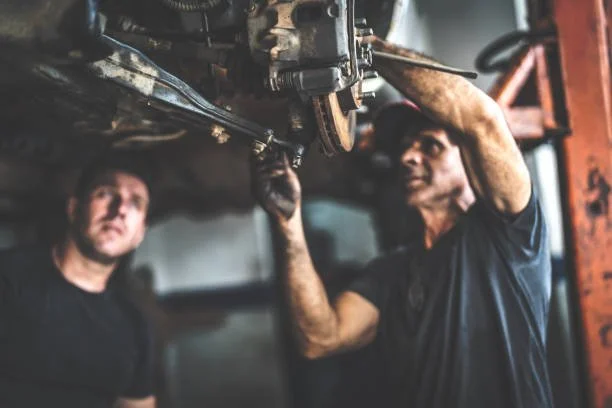 Two mechanics working under a car, one using tools to repair it in a garage.