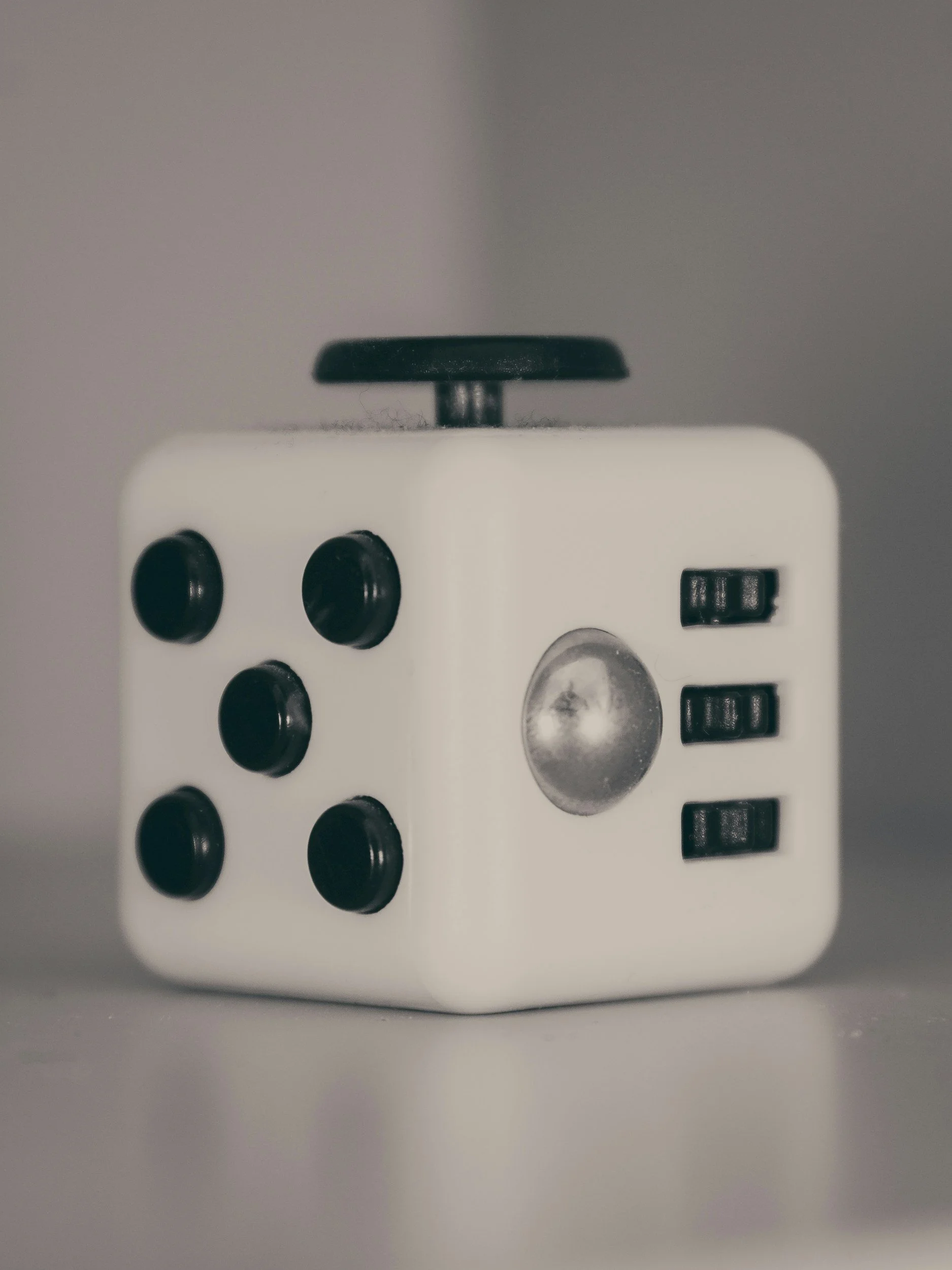 A fidget toy that is a white cube-shaped puzzle box with black buttons and a switch on the side, with a black dial on top and a small circular transparent window.