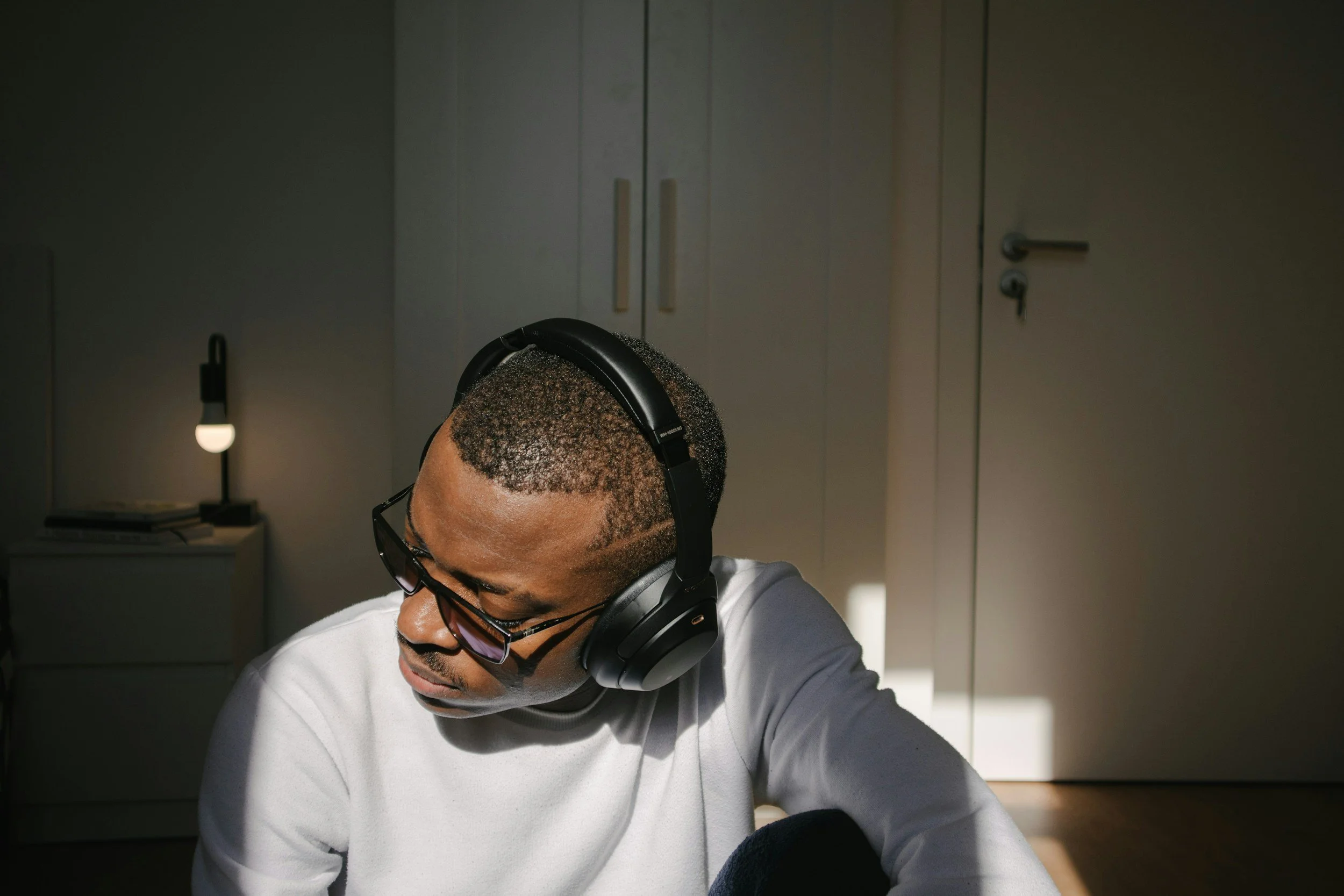 A man wearing glasses and headphones sitting in a dimly lit room with a closed cabinet in the background, listening to the Safe and Sound Protocol (SSP).