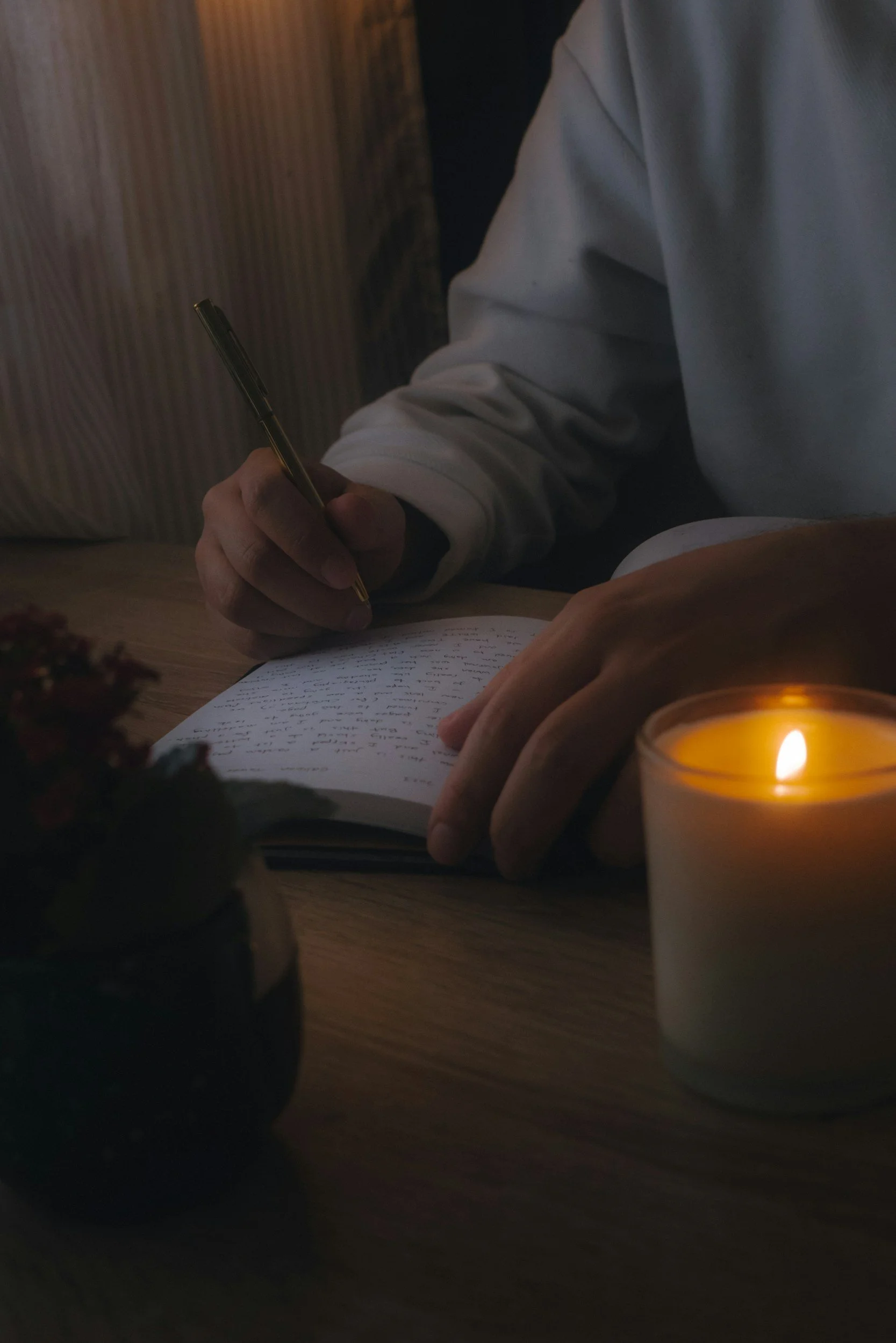 A person journaling self-reflections in a notebook with a pen, sitting at a table illuminated by a lit candle, with a blurred flowerpot in the foreground.