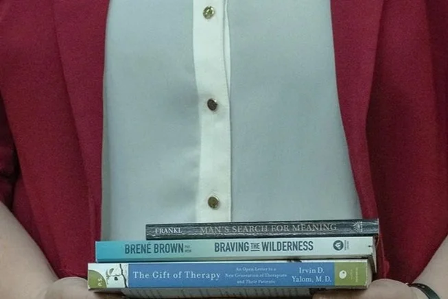 A person holding a stack of three mental health therapy books in front of their torso, wearing a white shirt and a red blazer. The books include the authors Brené Brown, existential approaches of Viktor Frankl and Irvin Yalom.