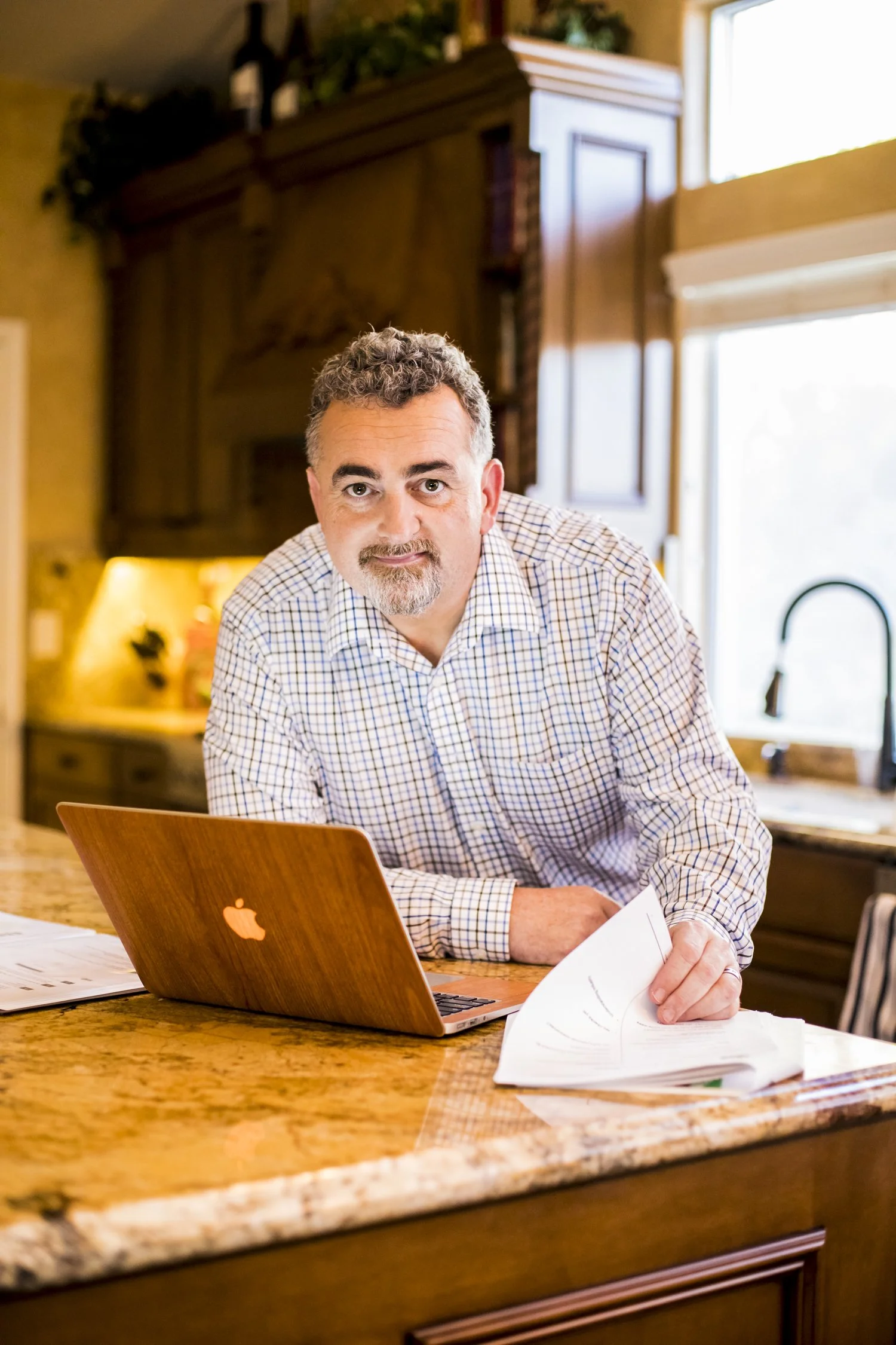 A middle-aged man with curly hair and a beard in a checked shirt, leaning over a kitchen countertop with a wooden MacBook and papers, in a cozy, well-lit kitchen.
