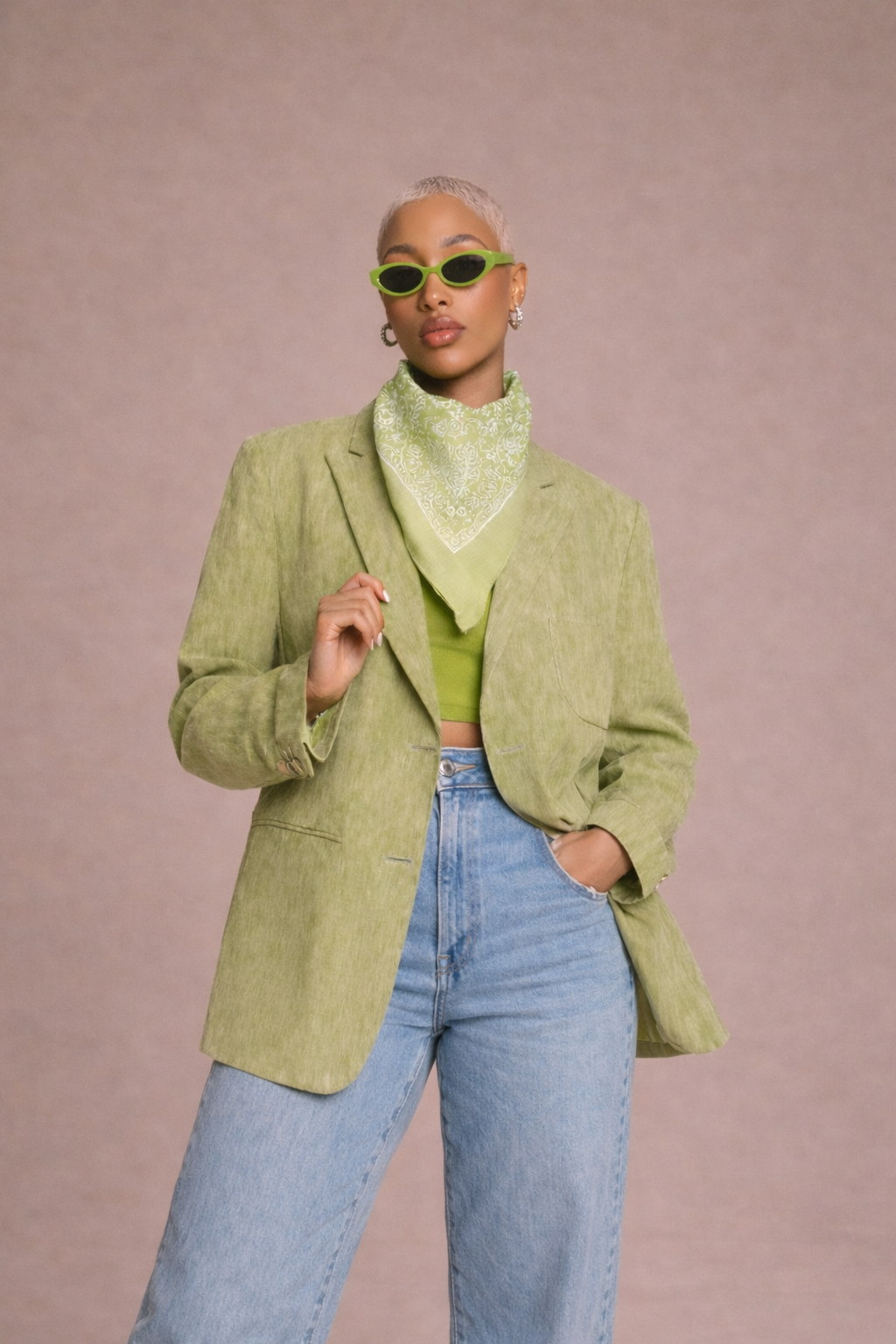 A woman with short blonde hair wearing green sunglasses, a green bandana, a green crop top, a green blazer, and light blue jeans, standing against a plain background.