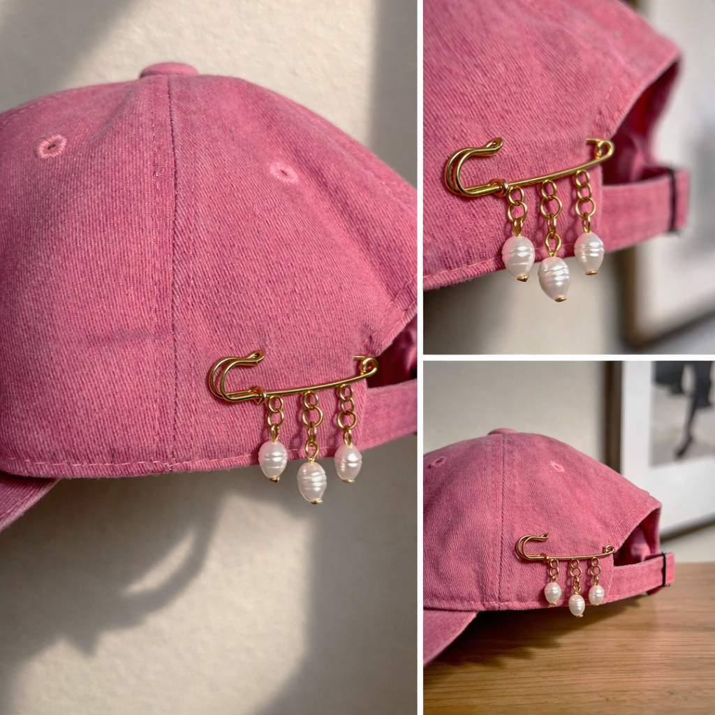 A pink baseball cap with a gold safety pin pin and pearl charms attached. The cap is shown from different angles, highlighting the pin and dangling pearl embellishments.