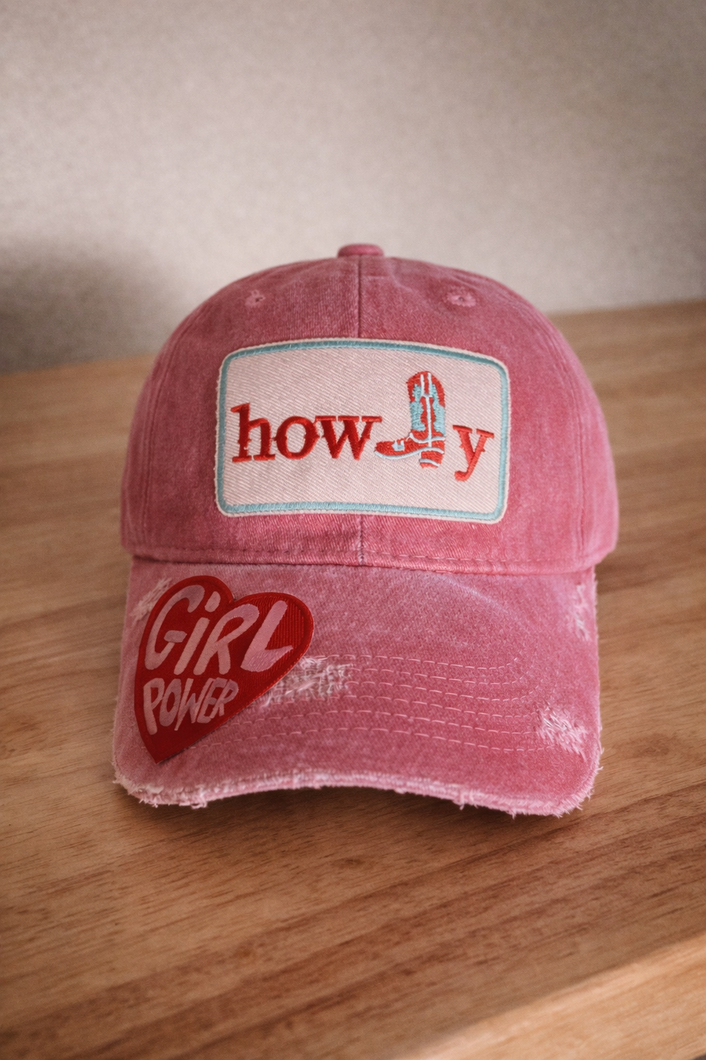 Pink distressed baseball cap with a patch that says 'howdy' featuring a cowboy boot graphic and a red heart patch labeled 'GIRL POWER' on the brim, placed on a wooden surface.