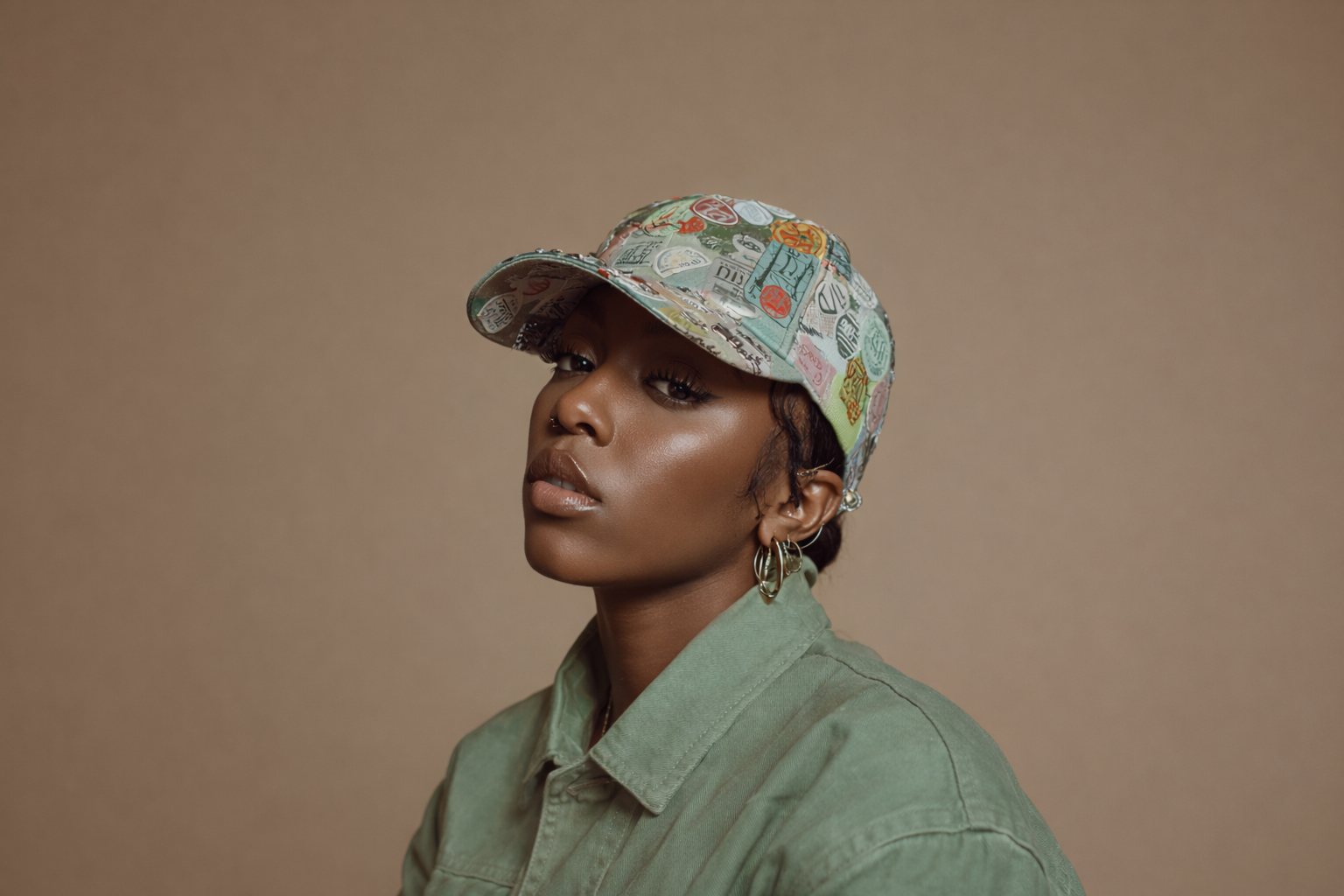 A woman wearing a colorful patterned baseball cap, earrings, and a green jacket against a beige background.