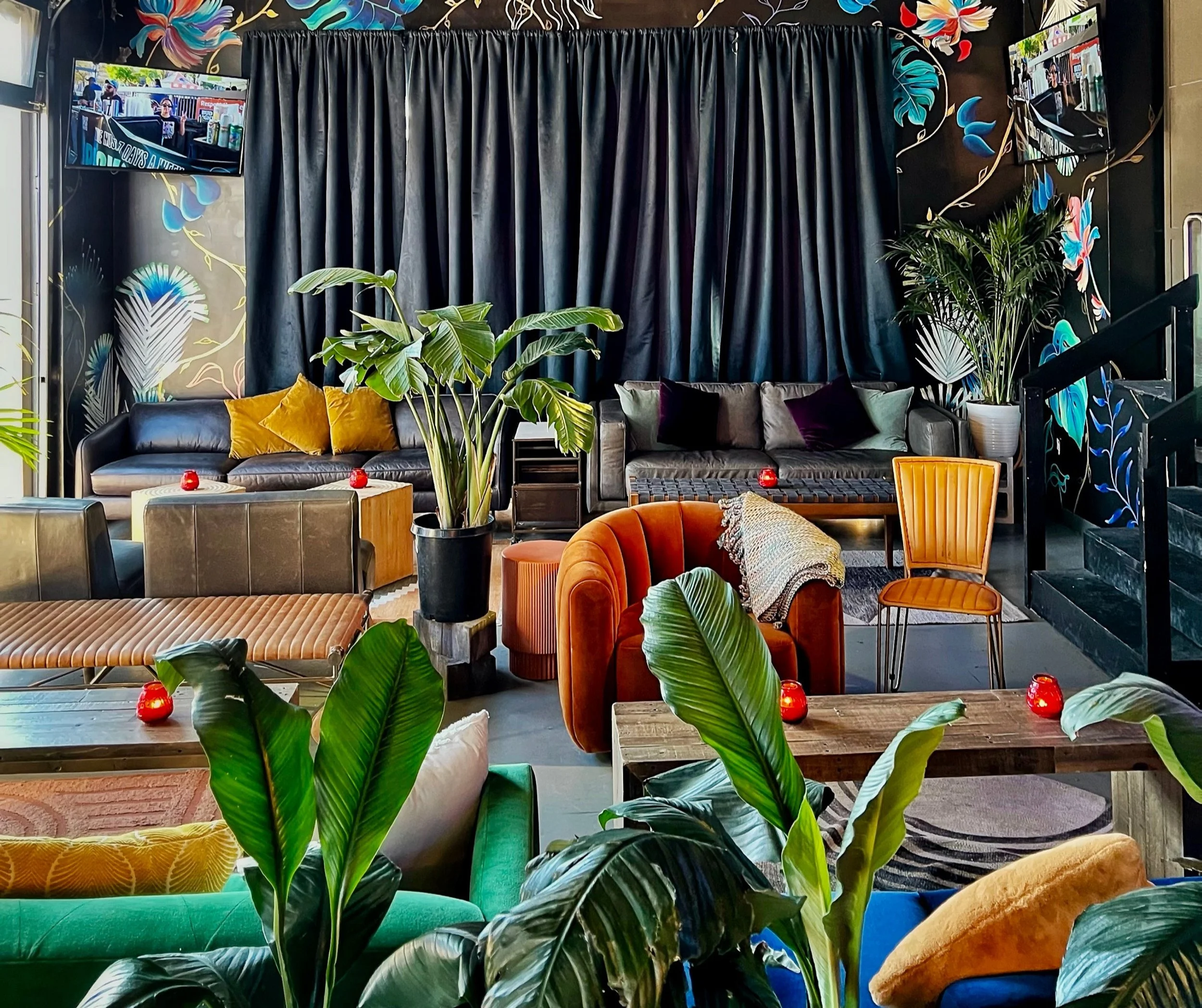 A brightly decorated lounge area with various colorful chairs, sofas, and potted plants, featuring a dark floral mural on the wall, black curtains, and two wall-mounted screens.