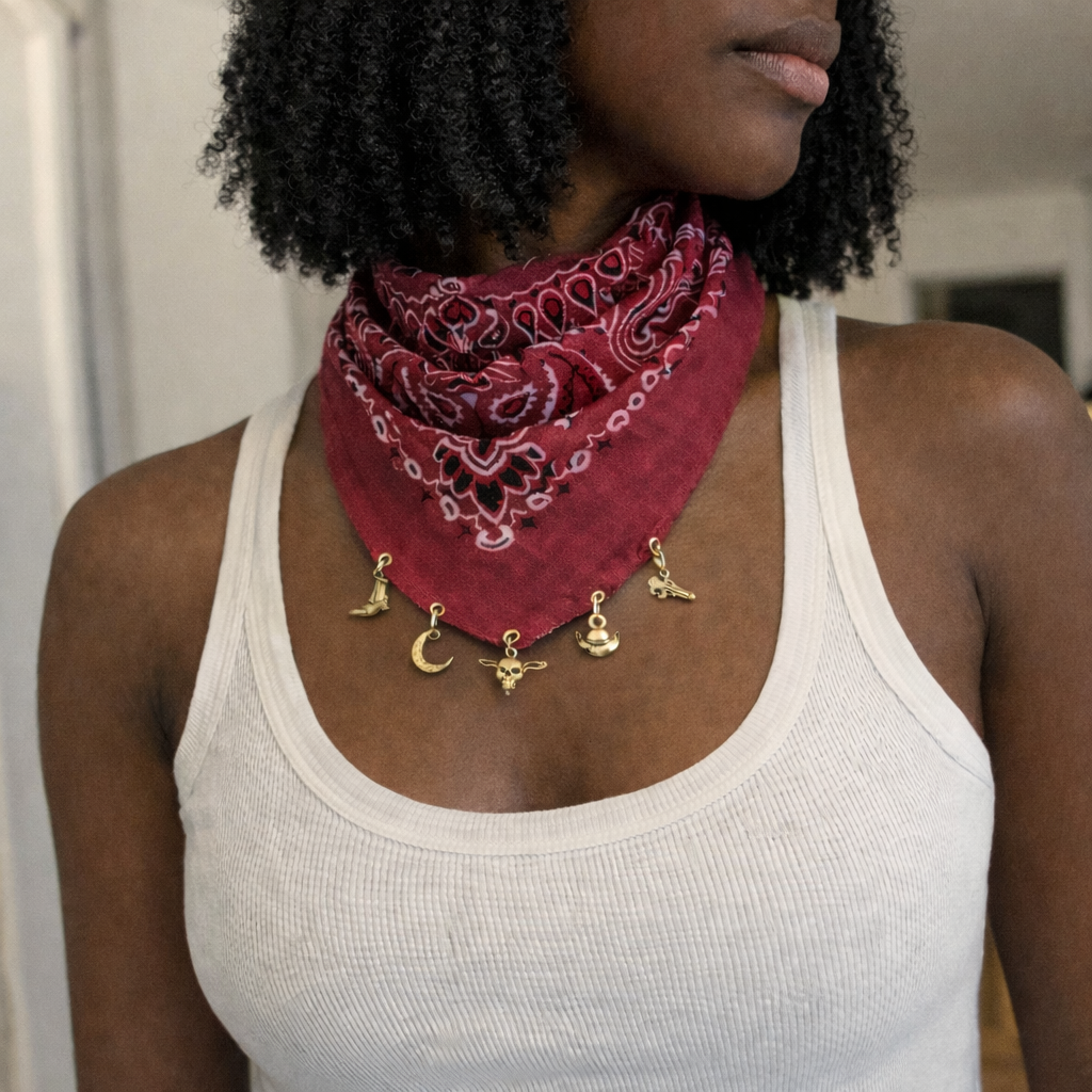 A woman wearing a red bandana around her neck with gold charms, including a moon, star, skull, horse, and handshake, over a white tank top.