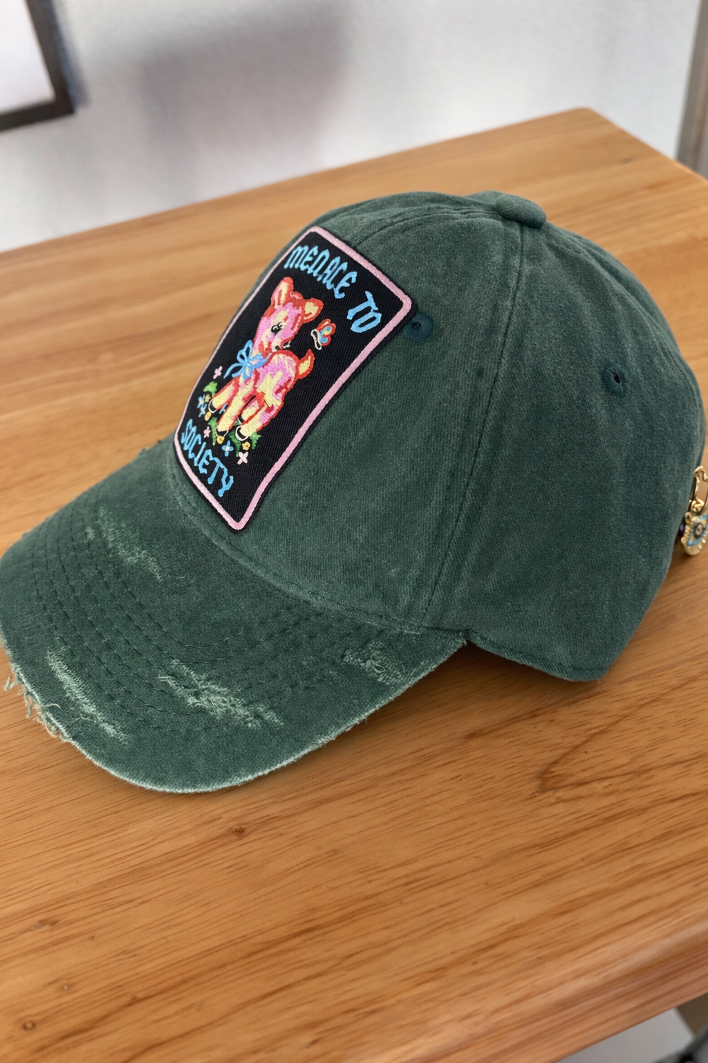 A green baseball cap with a colorful embroidered patch that features a cartoon pig, flowers, and the words 'Medicate to Save 21' on a wooden surface.