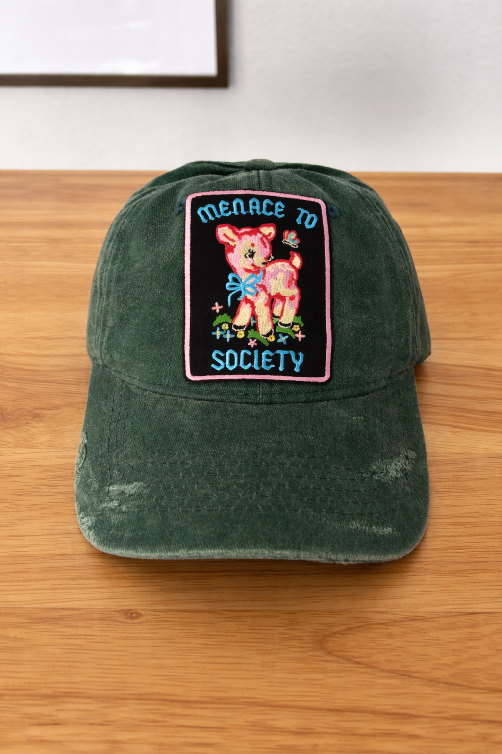 Green distressed baseball cap with a colorful patch featuring a cartoon deer and the text 'Menace to Society'.