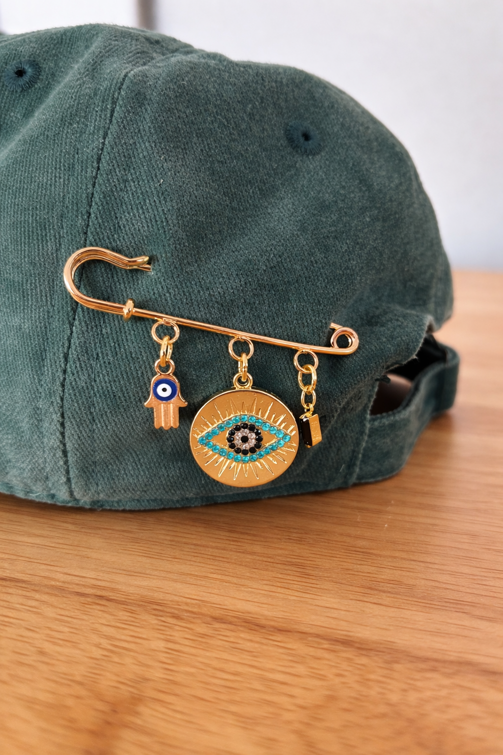 Green baseball cap with gold safety pin brooch featuring four charms: a hand with an eye symbol, a large eye with turquoise accents, a small rectangular charm, and an eye symbol.