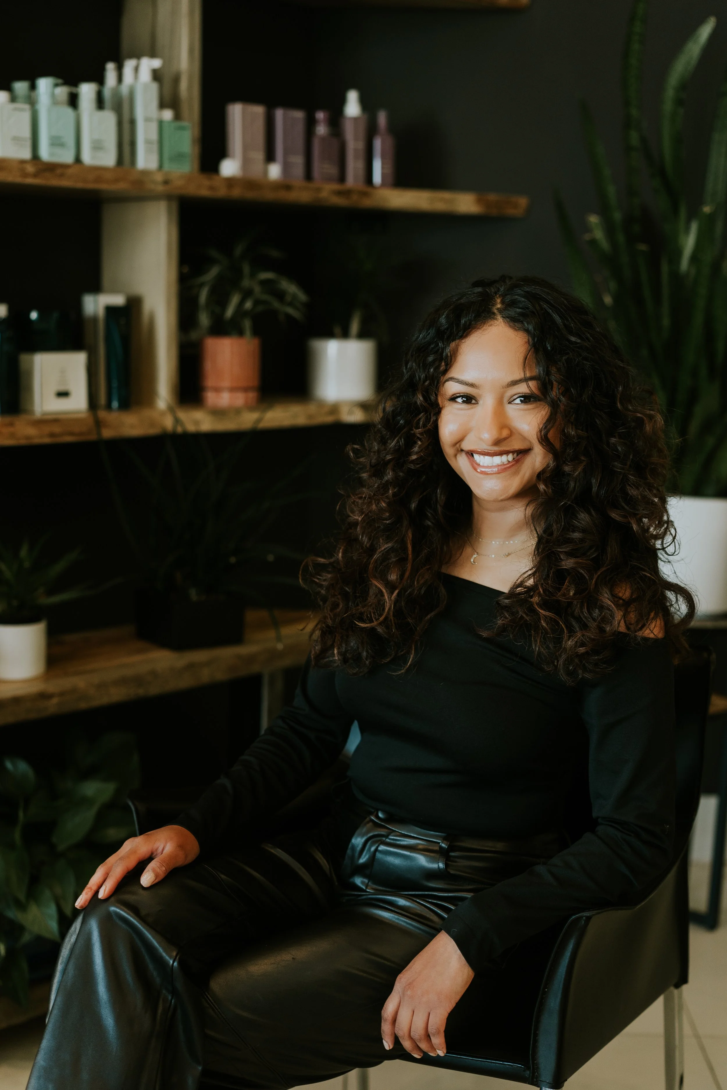 Mujer-líder-segura sonriendo sentada en una silla de oficina en un ambiente con plantas y estantes de madera con productos y libros.