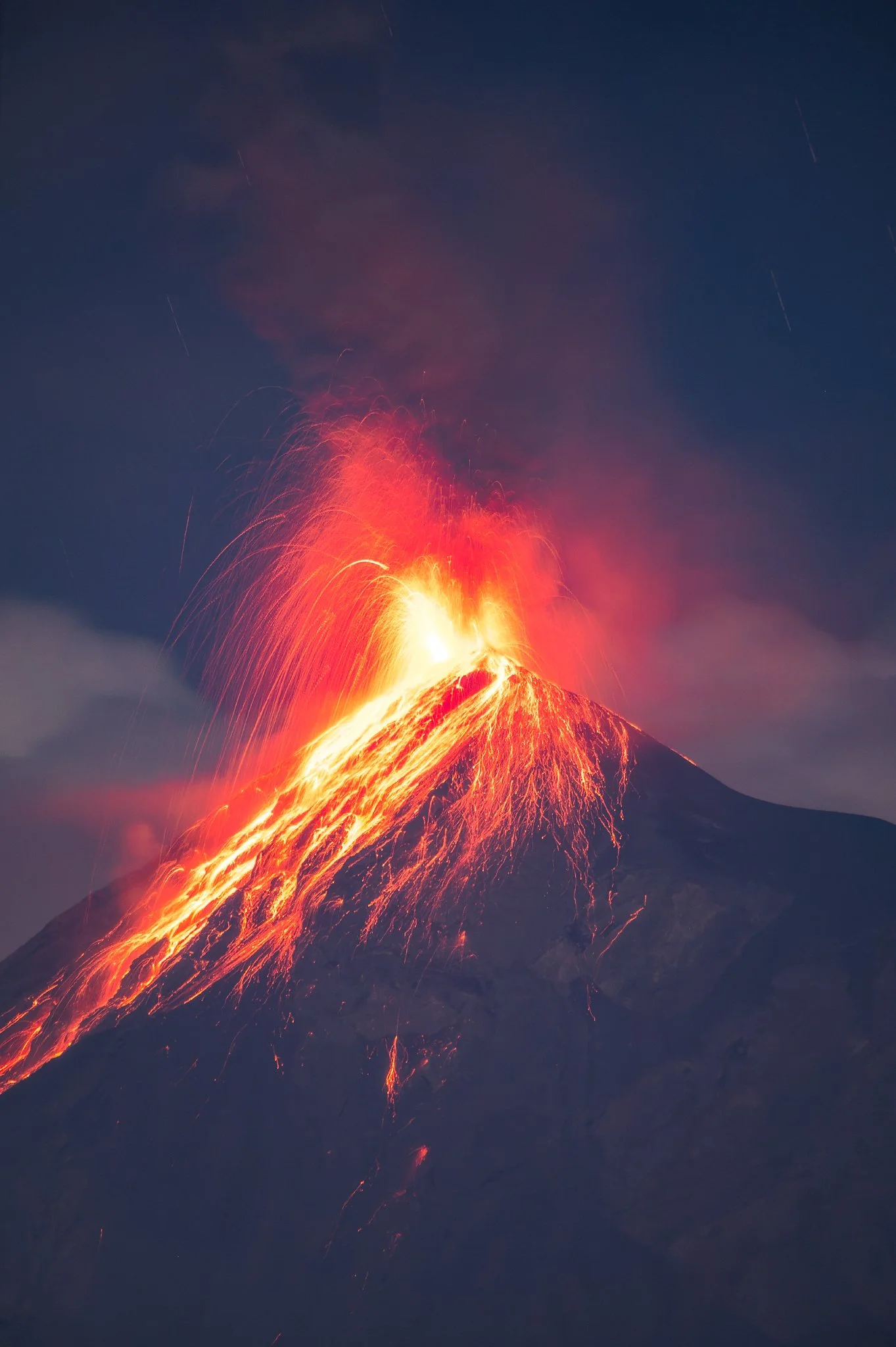 Eruption of a volcano at night with bright red lava flowing down the sides and ash and smoke rising from the crater.