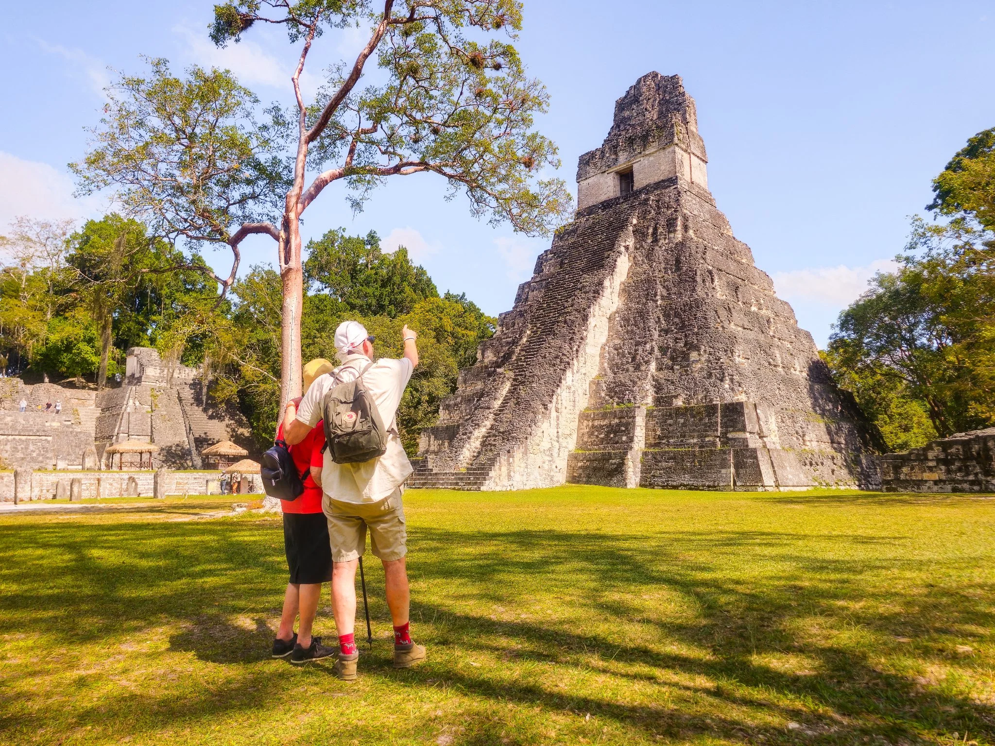 Two tourists, one wearing a white hat and beige shirt, and the other in a red shirt, stand on grass pointing towards an ancient pyramid surrounded by trees at an archaeological site.