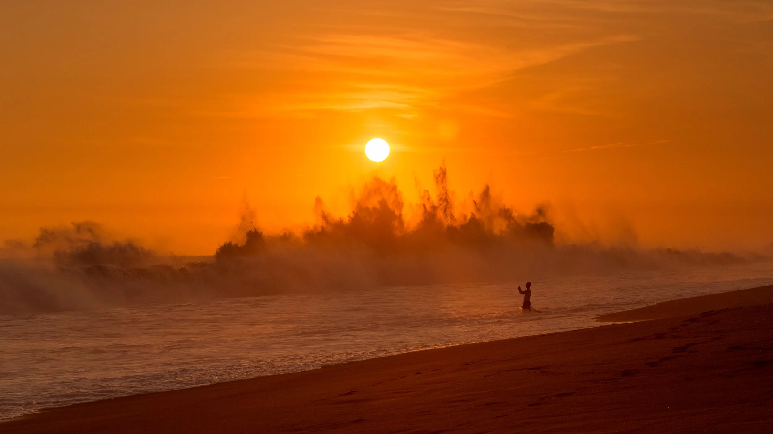 Person standing in shallow ocean water at sunset with a large, bright sun in the sky, orange and yellow hues, gentle waves, and mist in the air.